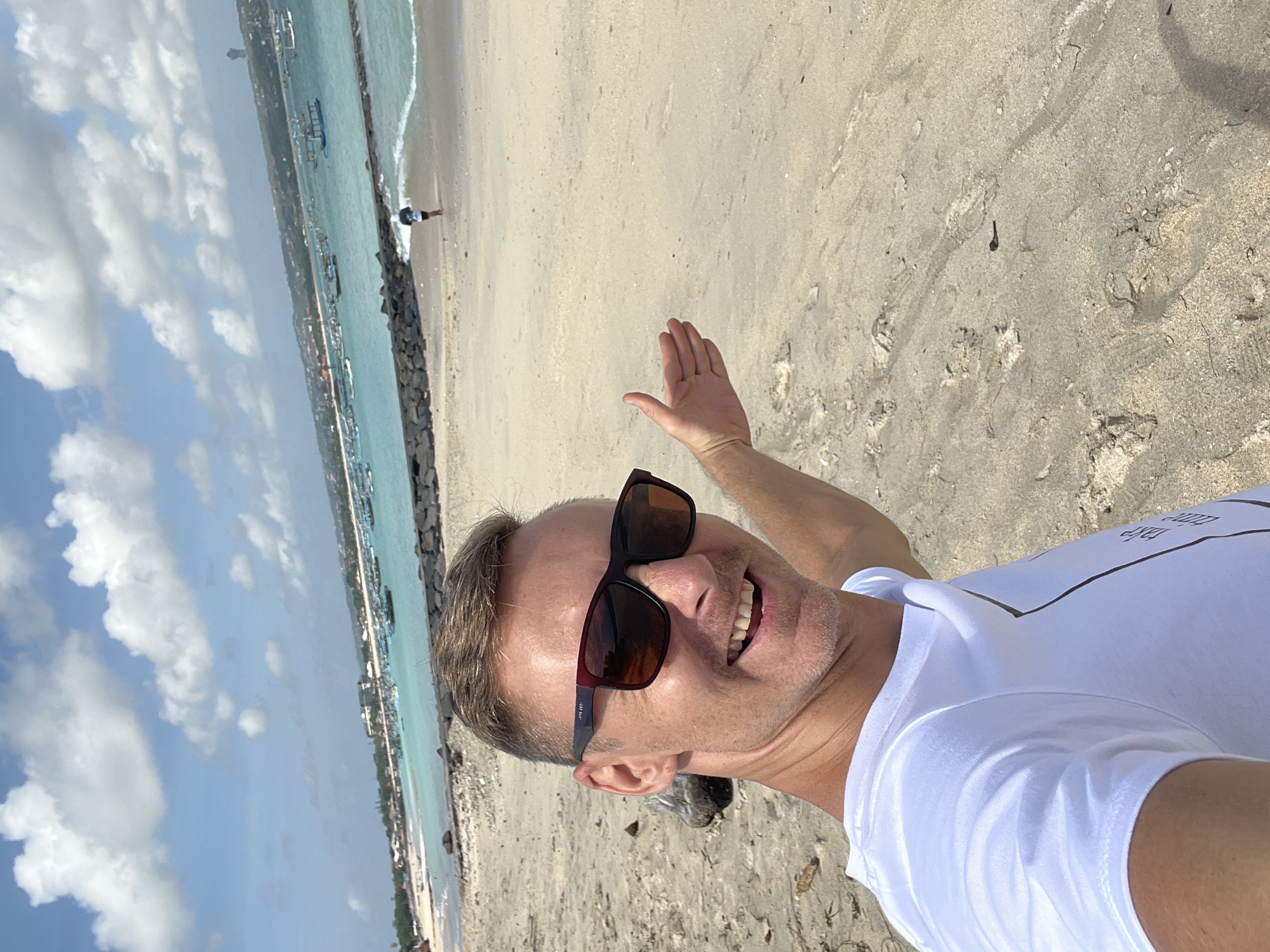 A man in sunglasses and a white t-shirt takes a selfie on a sandy beach in Kuta, Indonesia. He is smiling and gesturing with his right hand towards the ocean, which is filled with many boats and has a breakwater along the coast. In the background, a person is seen running along the shoreline. The sky is blue with scattered white clouds.