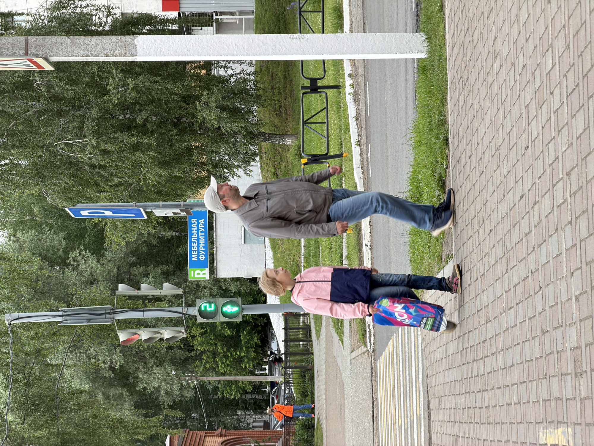An outdoor street scene in the city of Chaikovsky, Russia, depicts two pedestrians on a paved sidewalk next to a road, under an overcast sky.
A young girl, approximately 8-12 years old, wearing a pink and navy blue hooded jacket, blue jeans, and multi-colored sneakers, walks on the sidewalk carrying a blue shopping bag with "ZON" visible on it. Her head is turned slightly to the right, looking forward. Directly behind and to the right of her, an adult male, likely in his late 30s to 50s, walks in the same direction. He wears a light grey baseball cap, a light brown/grey leather-like jacket, blue jeans, and dark sneakers. He also looks slightly to his right.
To the left of the pedestrians, a striped pedestrian crosswalk is visible on the asphalt road. A traffic light pole stands prominently in the mid-ground. One traffic light facing the viewer displays a green walking person symbol with the number "2" illuminated, indicating 2 seconds remaining for pedestrians to cross. Another traffic light on the same pole, oriented away from the camera, shows a red light.
In the background, lush green trees with thin trunks (possibly birch) line the street. A light-colored building in the mid-ground features a green rectangular sign with "R" and the Cyrillic text "МЕБЕЛЬНАЯ ФУРНИТУРА" (Furniture Fittings). To the far left, a red brick building with arched windows is partially visible. Further back on the left, an adult in a bright orange jacket is visible behind a black metal fence, appearing to be holding a phone or camera. Parked cars are visible behind this fence. A prominent white pole stands on the far right of the image. Grass and other vegetation are visible along the roadside and in the distant background.