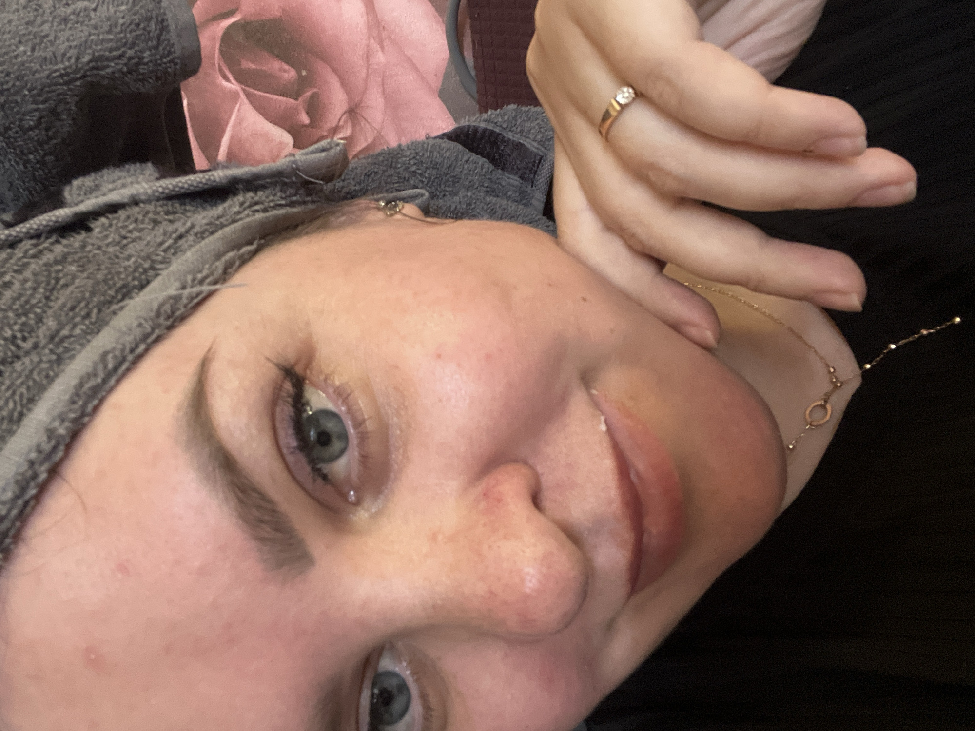 A close-up, tilted shot focuses on a fair-skinned woman with blue eyes, freckles on her nose, and well-defined eyebrows. She is wearing a gray towel wrapped around her head. Her hand, adorned with a gold ring featuring a small stone, is positioned near her face. A gold chain necklace with small circular elements is visible. The background includes a rose-patterned surface and a dark, textured fabric, possibly clothing. The location of Noviy, Russia is not discernible from the image.