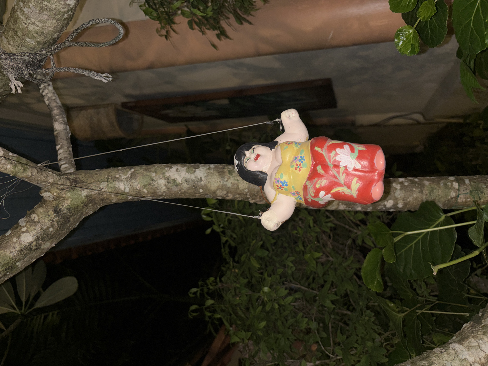 In Chalong, Thailand, a ceramic figurine of a smiling, plump woman with black hair, wearing a yellow floral halter top and red pants with white flowers, hangs from a tree branch by thin white strings. The figurine is suspended in the foreground. To the right, a thick rope is tied to another tree branch, with loose ends hanging. Behind the figurine and rope, lush green foliage is visible, with dark shadows suggesting nighttime. In the background, a building with a textured, light-colored wall and a dark roof can be seen, with a framed picture hanging on the wall. The scene is dimly lit, with artificial light illuminating the figurine and the immediate surroundings.