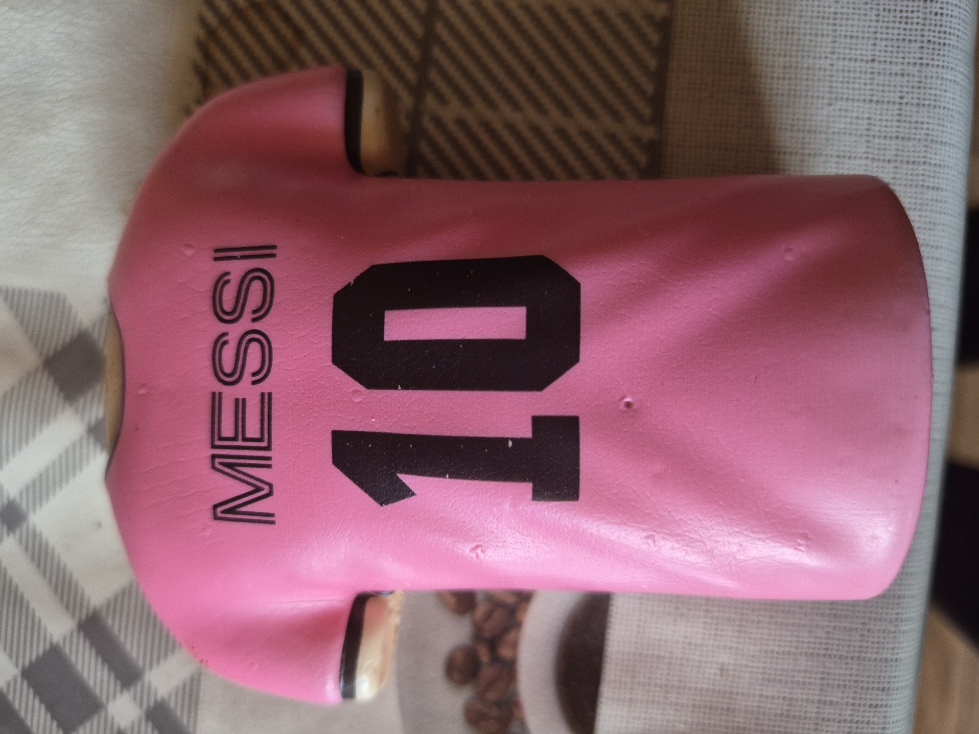 The image features a pink object shaped like a football jersey, adorned with the name "MESSI" printed vertically in black capital letters on the left side and the number "10" in large black numerals on the right. The jersey's surface is slightly uneven and glossy, suggesting it might be an iced cookie, a molded decorative item, or made of a pliable material like fondant. Small indentations and textures are visible on its surface.
The jersey is positioned on a light-colored surface with a grey and white checkered pattern, likely a tablecloth or mat. In the background, particularly in the lower right, a blurred white object is visible, appearing to hold numerous brown coffee beans, and the dark rim of what seems to be a mug or bowl. Another patterned surface with diagonal stripes in shades of brown and black is partially visible in the upper background.
No people or activities are depicted; the image is a static close-up of the themed item. The setting is indoors. The time of day and weather cannot be determined. The visible text "MESSI" and "10" clearly indicate a tribute to the famous footballer, Lionel Messi. The context provided (Yerevan, Armenia) is not visually evident in the image itself.