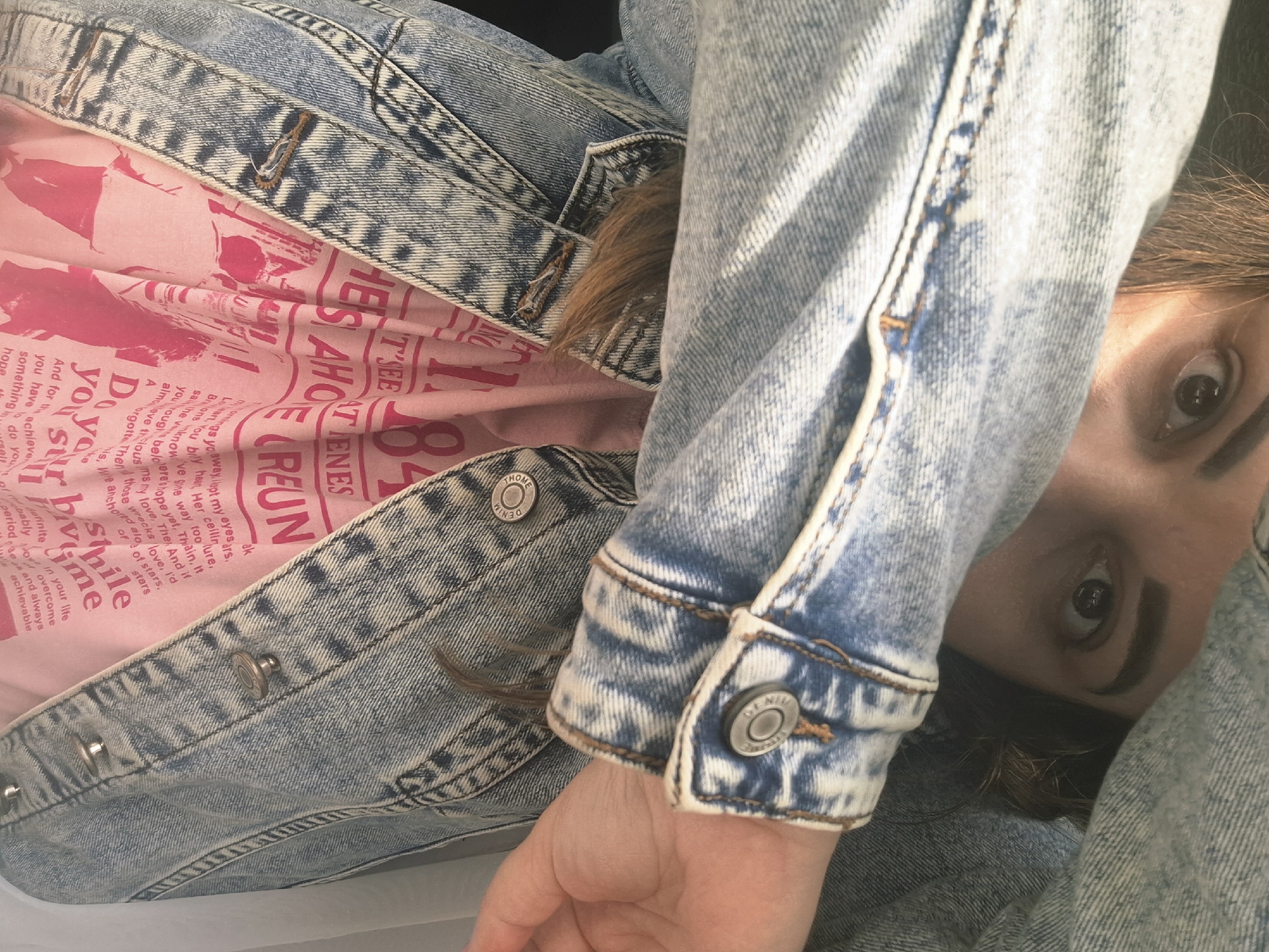 The image shows a person with brown eyes partially visible, framed by a light-wash denim jacket. The person is wearing a pink t-shirt with text printed on it, partially obscured by the jacket. The visible text includes phrases like "smile", "Do your thing," and the word "REUN". The jacket is heavily textured with a white, acid-wash pattern. The person's hand and part of their arm are also visible. There is no information in the image that suggests the location is in the city of Chaikovsky, Russia.
