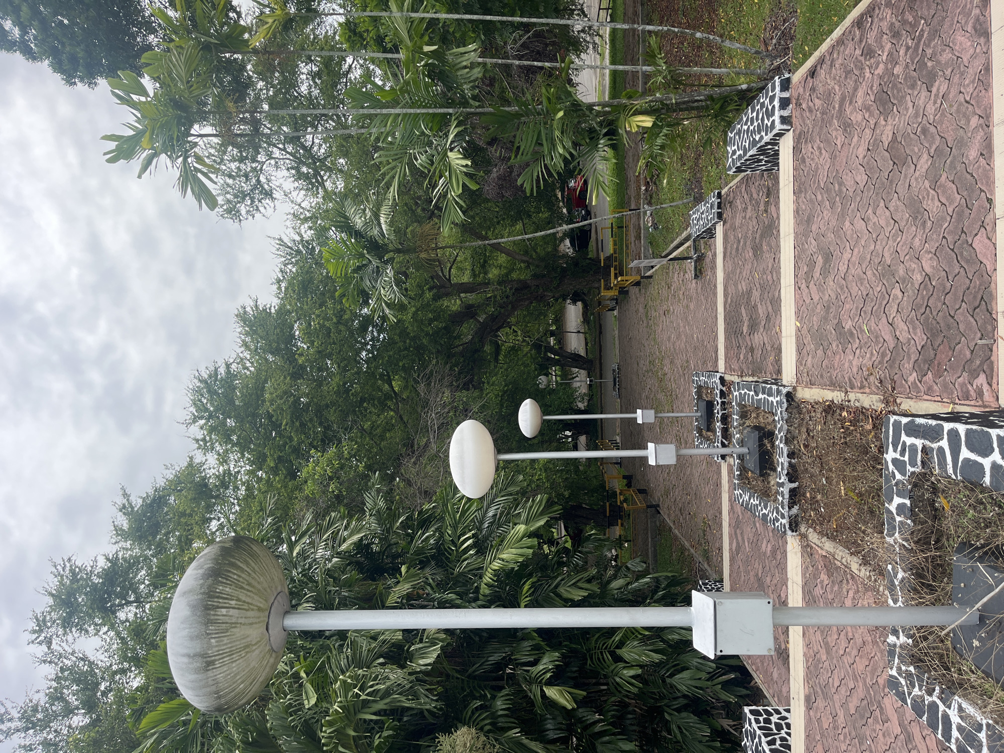 The image shows an outdoor scene in Lhokseumawe, Indonesia. Three spherical streetlights line a brick-paved pathway bordered by raised planters with a black and white "cow-spotted" design. Lush green trees and foliage form a backdrop. In the distance, a yellow scaffolding structure and a red vehicle are visible. The sky is overcast.