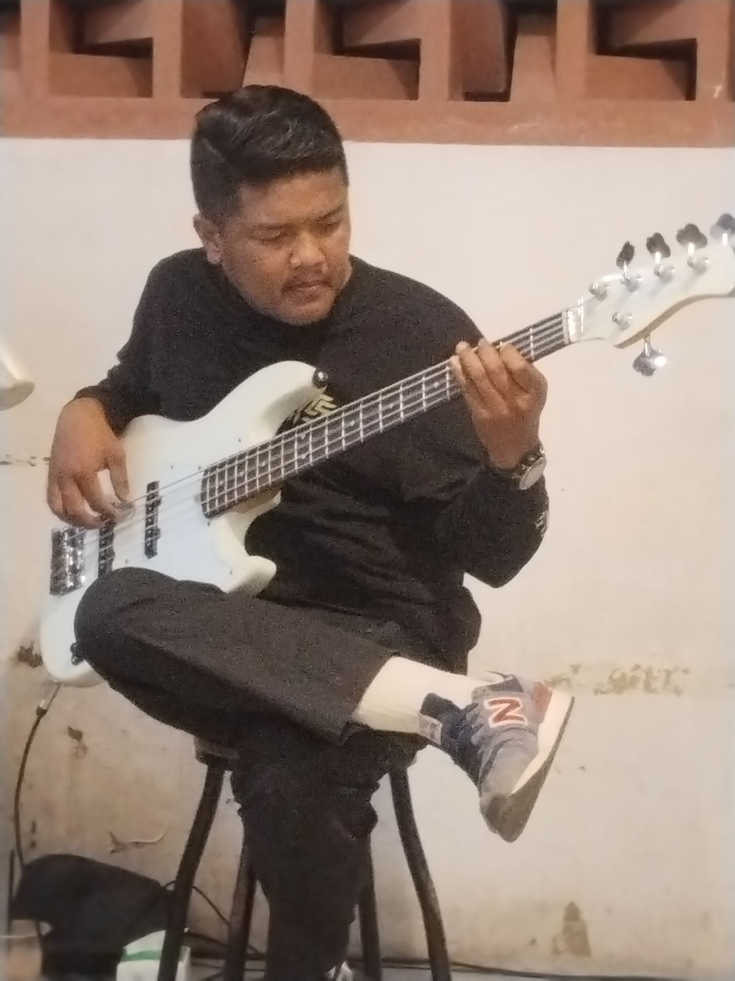 A man sits on a stool, playing a white electric bass guitar. He wears a black long-sleeved shirt, dark pants, white socks, and grey New Balance sneakers with a red "N" logo. His hair is short and dark, styled back. He is focused on playing the bass. The background is a plain wall with a decorative brick pattern near the top. A cable is visible near the base of the bass guitar. The location is Lhokseumawe, Indonesia.