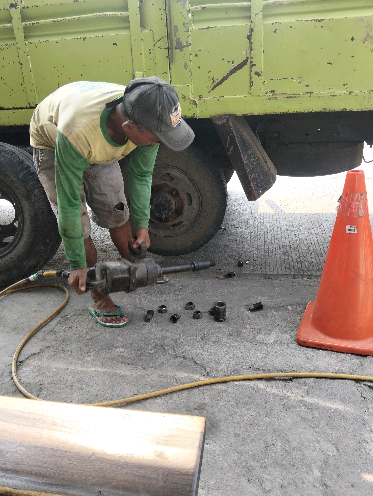 A man wearing a green long-sleeved shirt, grey shorts with a hole in the left knee, and a dark baseball cap is working on a truck. He is using a large, black pneumatic impact wrench, holding it with both hands. The wrench is connected to a yellow hose, likely for air supply. The man is wearing green flip-flops. Several metal sockets and nuts are scattered on the ground around him. The truck is large and green, with some signs of wear and tear. It is parked on a concrete surface, possibly a roadside or workshop area. An orange traffic cone is visible to the right of the frame, and a portion of a wooden beam is in the lower left corner. The scene suggests a truck maintenance or repair activity in an outdoor urban environment.