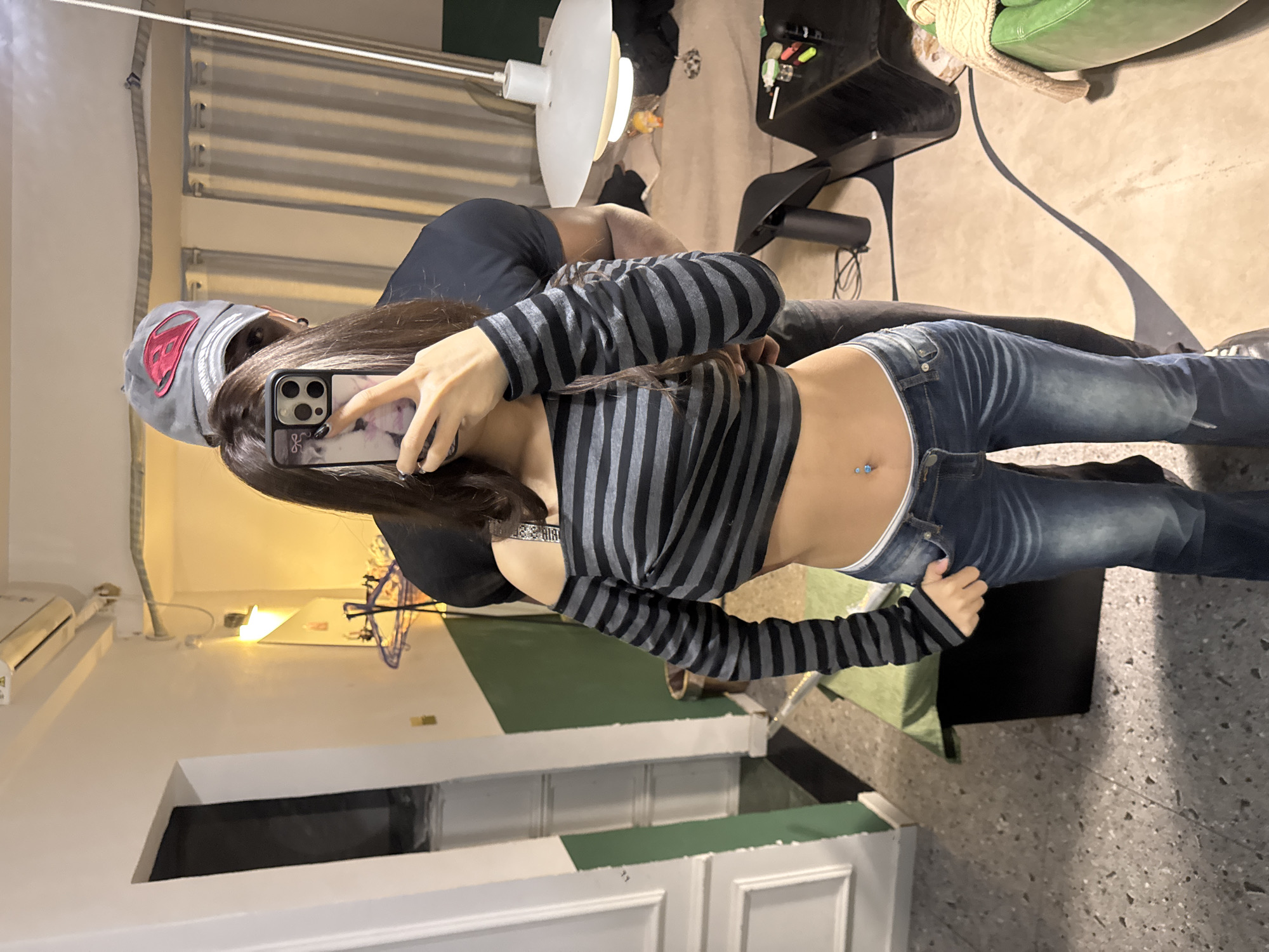 The scene depicts two individuals indoors in Liangjiang, China. In the foreground, a female subject stands facing the camera, her face obscured by a silver-colored smartphone held in her right hand. She wears a black and grey horizontal striped, off-the-shoulder, long-sleeved crop top, exposing her midriff, which features a blue-gemmed belly button piercing. She is dressed in low-rise blue denim jeans, revealing a white waistband. Behind her, a male subject is positioned, facing away from the camera, with his arms around the female's waist. He wears a grey baseball cap with a red logo, dark sunglasses, and a dark-colored t-shirt. A dark ear stud is visible on his left ear.
The environment appears to be a residential or common area. The walls are primarily white, with a dark green wainscoting on the lower left section. A white doorway with molded trim is visible on the left. The floor is a speckled, light grey terrazzo or concrete. Overhead, an air conditioning unit is mounted on the left wall, and a large, white, tiered pendant light fixture hangs from the ceiling on the right. A wall-mounted light fixture with a yellowish glow is present on the left wall. In the background, vertically striped light-colored curtains cover an opening or window. Furniture visible includes a dark-colored, rectangular table or console with a green fabric cover on the left, a dark coffee table with small items on it (e.g., markers, a lighter) on the right, and a green armchair with a cream-colored knit blanket draped over it on the far right. A light-colored sofa or bed is partially visible in the distant background. Shadows are cast across the floor from the subjects and furniture.