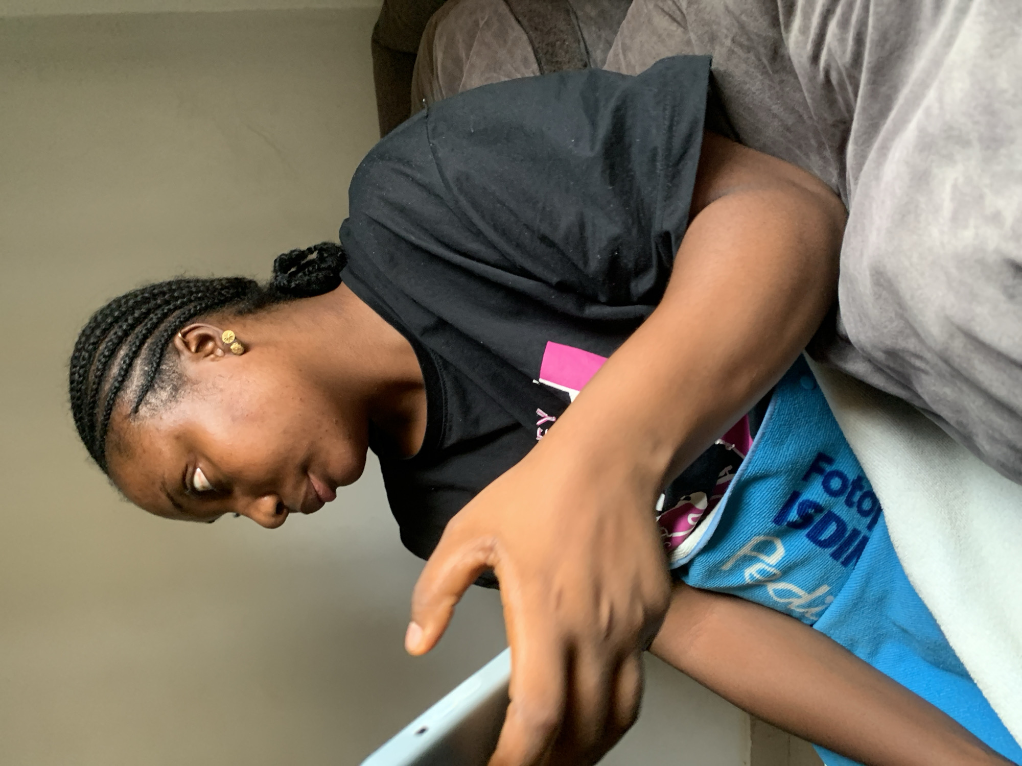 A medium shot of a dark-skinned woman with braided hair, wearing a black t-shirt, seated on a grey couch. The woman is looking down and towards the left, and her right hand is extended, holding what appears to be a tablet. A blue towel with white lettering is draped across her lap. The background is a plain wall. The location cannot be determined to be Abuja, Nigeria, as there are no discernible clues.