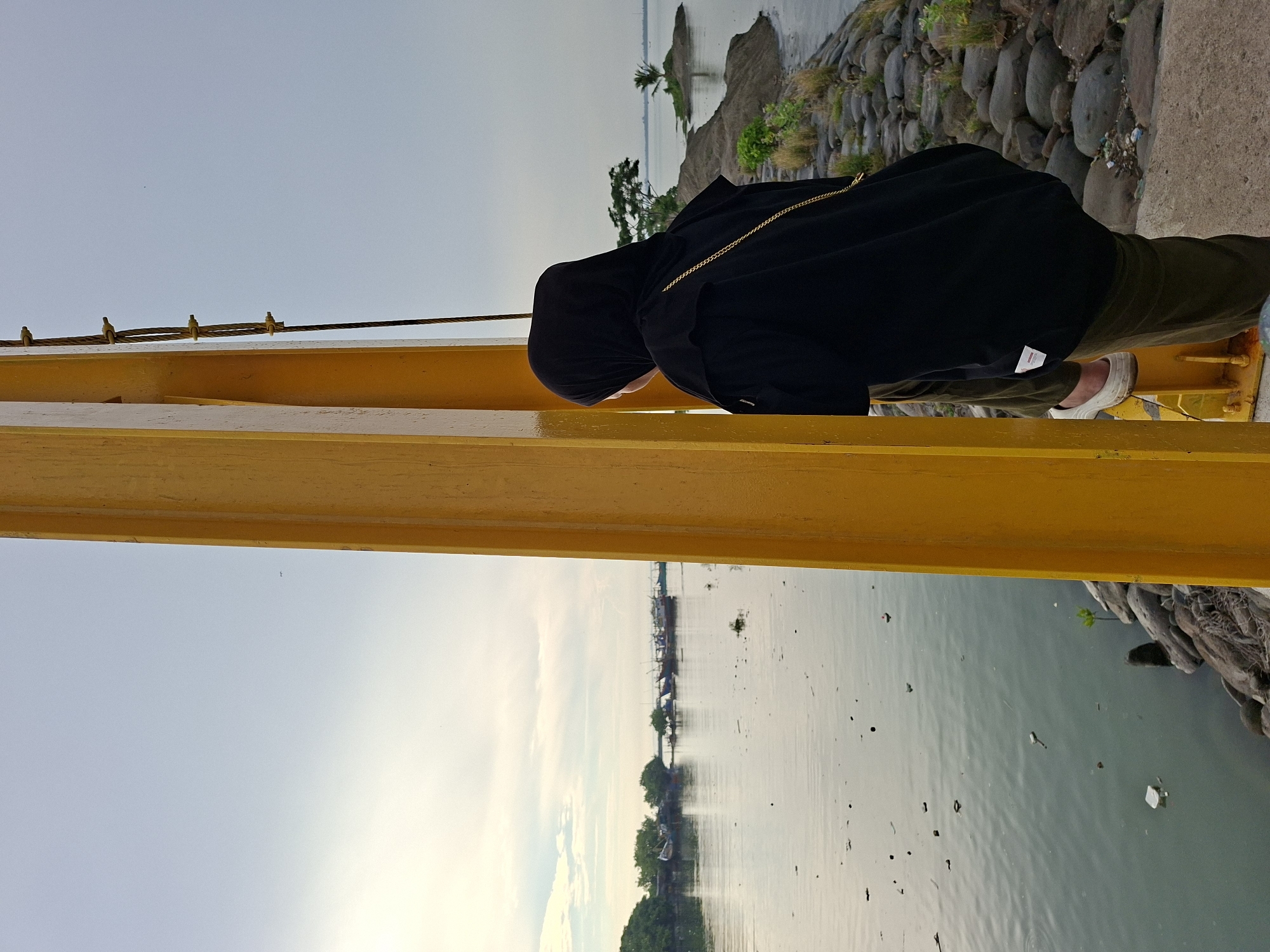 The image captures a woman wearing a black hijab and a black oversized shirt, standing by a yellow structure, likely a bridge. She is looking out at a body of water, which appears to be a harbor or bay. In the background, several boats are docked, and a small island with a single tree is visible. The shoreline is lined with rocks and some vegetation. The sky is overcast with some clouds. The scene is likely the Lhokseumawe Reservoir or a similar coastal area in Lhokseumawe, Indonesia, given the presence of boats and the general landscape. The water contains visible debris, suggesting a potentially polluted environment. The woman's posture suggests contemplation or observation of the seascape.