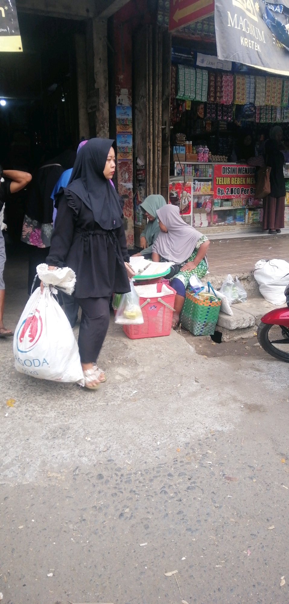 In a street market scene in Ajibarang, Indonesia, a woman wearing a black hijab and black tunic walks to the left carrying a large white "GODA 50 KG" bag and a plastic bag. Two women are seated on the ground near a stall displaying various products, one wearing a light green hijab and the other a grey hijab. A red basket and a green basket are placed on the ground. The stall behind them is stocked with goods including bottled drinks, packaged food items, and eggs. A red motorcycle is partially visible on the right side of the image. The ground is paved with concrete and appears weathered. The overall environment suggests a bustling local market.