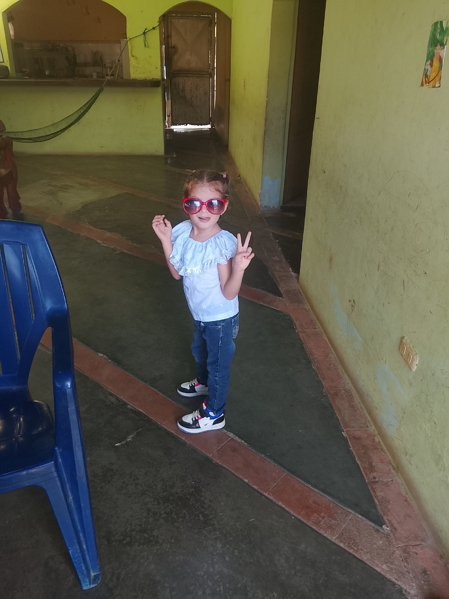 A young girl with pigtails and wearing red sunglasses, a light blue blouse, blue jeans, and black and white sneakers, stands indoors. She is making a peace sign with both hands. To the left of the girl is a blue plastic chair. The indoor space has yellowish-green walls and a floor with a dark green surface bordered by a red-brown trim. In the background is a doorway and a hammock hangs indoors. The image was taken in Altagracia, Venezuela.
