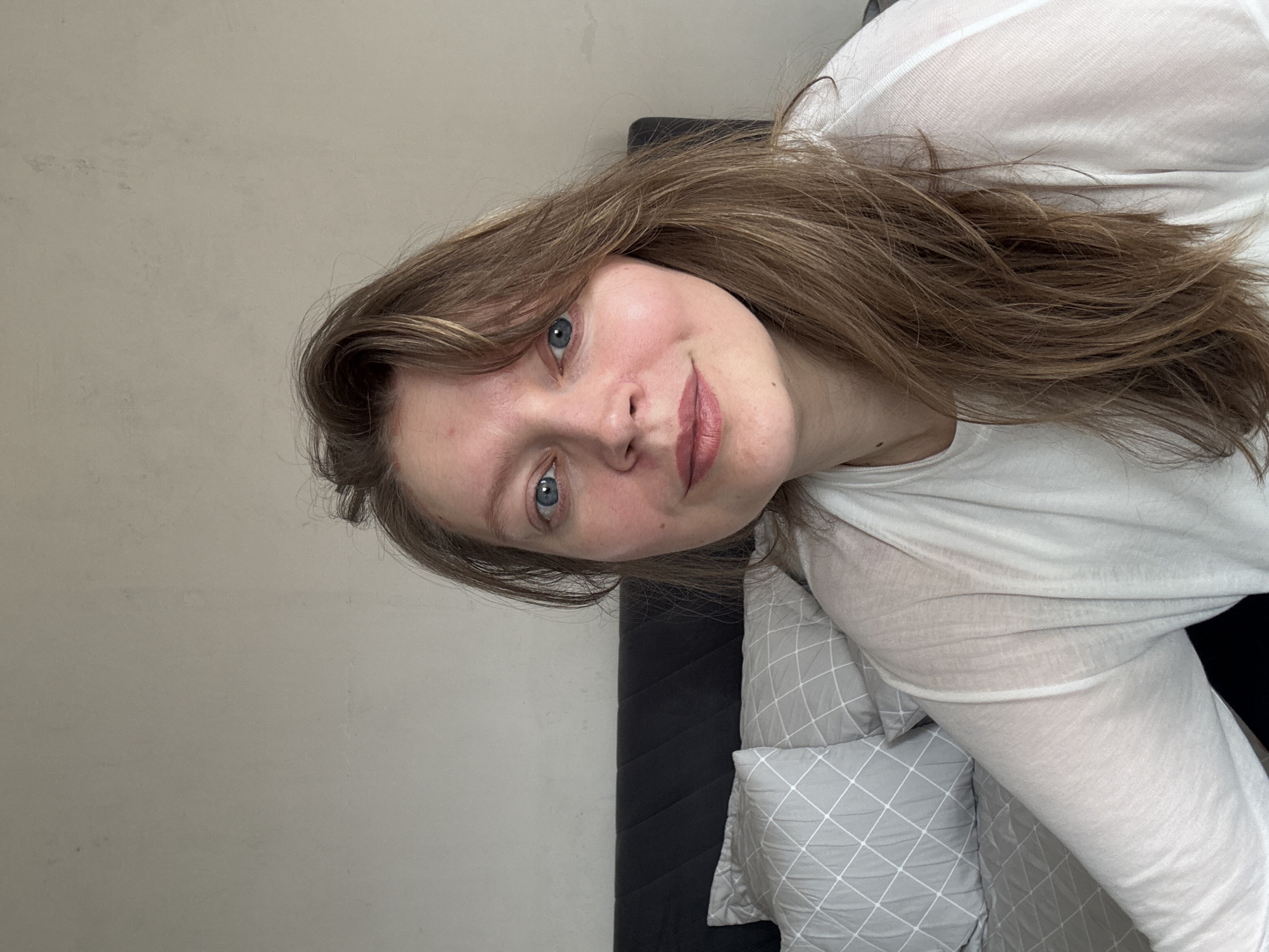 A woman with light brown hair and blue eyes is posing for a picture. She is wearing a white, semi-sheer shirt and is positioned against a gray wall and a black headboard. There are gray pillows with a white diamond pattern visible behind her. The image does not contain sufficient information to determine her exact location in Izhevsk, Russia, or any actions, interactions, or further environmental details.