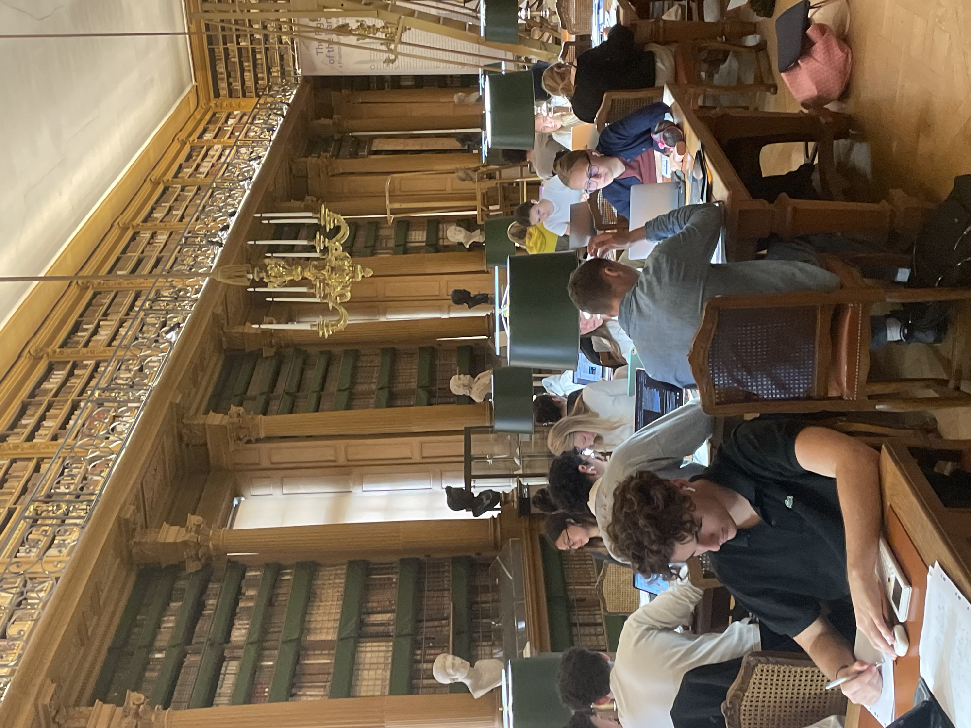 The image depicts the interior of the Bibliothèque Sainte-Geneviève, a public library located in the Latin Quarter of Paris, France. The scene is set in a grand reading room characterized by high ceilings, ornate wooden bookshelves filled with numerous books, and classical architectural elements such as columns and arches. Natural light streams in from large windows.
Several individuals, appearing to be students, are seated at long wooden tables, engaged in various activities related to their studies. Most are focused on laptops, with some writing or taking notes. One person in the foreground, a young male wearing a black polo shirt, is actively writing on a notepad with a pen, with a calculator and other papers on the table in front of him. Another person, a young female in the foreground, is wearing glasses and looking at a laptop.
The atmosphere appears quiet and studious, typical of a library environment. The presence of students, books, and study materials suggests an academic setting. The overall impression is one of intellectual pursuit within a historically rich and architecturally significant space.