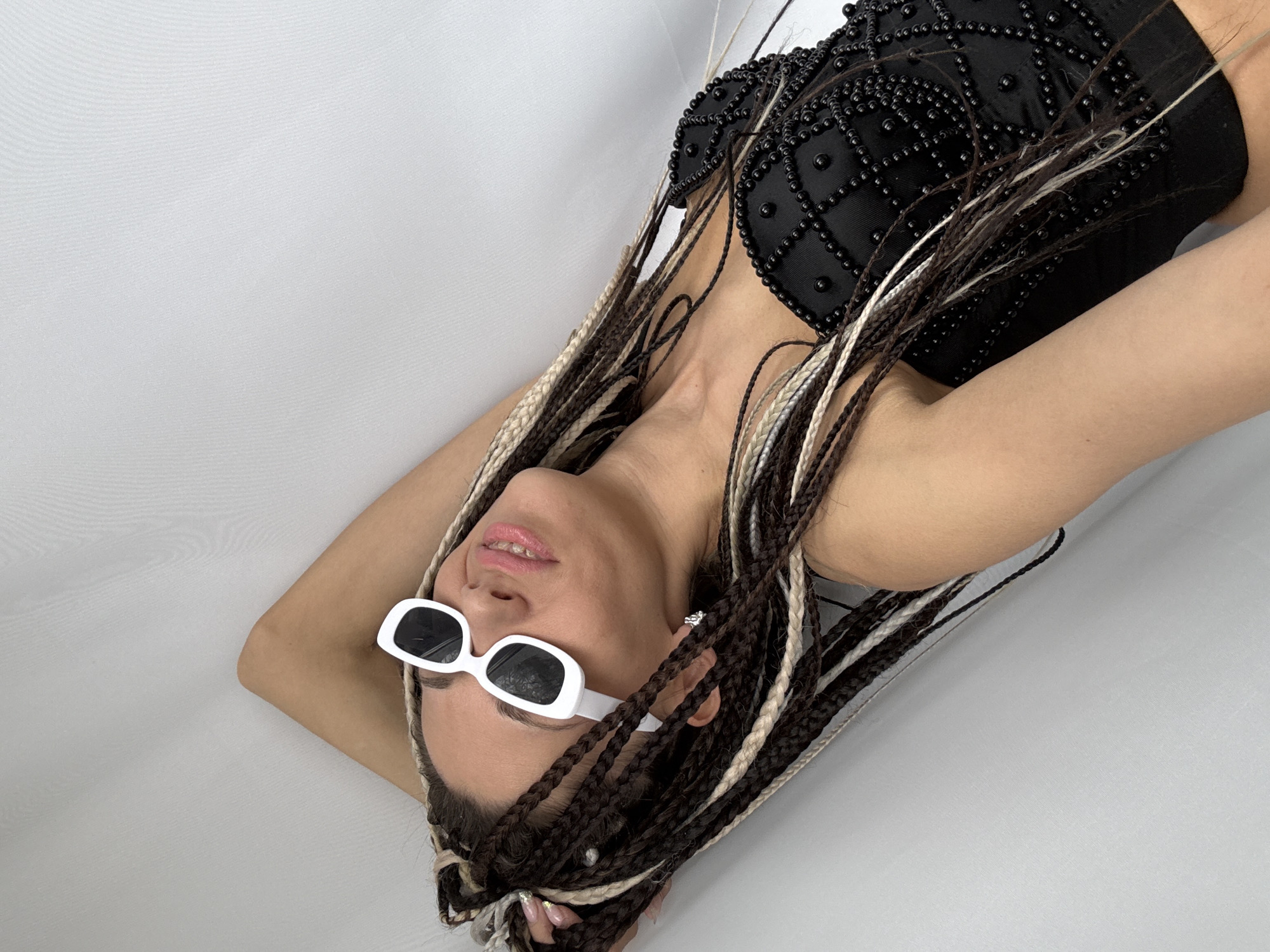 A woman with braided hair is lying on a white surface. She is wearing white sunglasses and a black top with embellishments. The location cannot be determined to be Rusanovo, Russia, based on the provided image.
