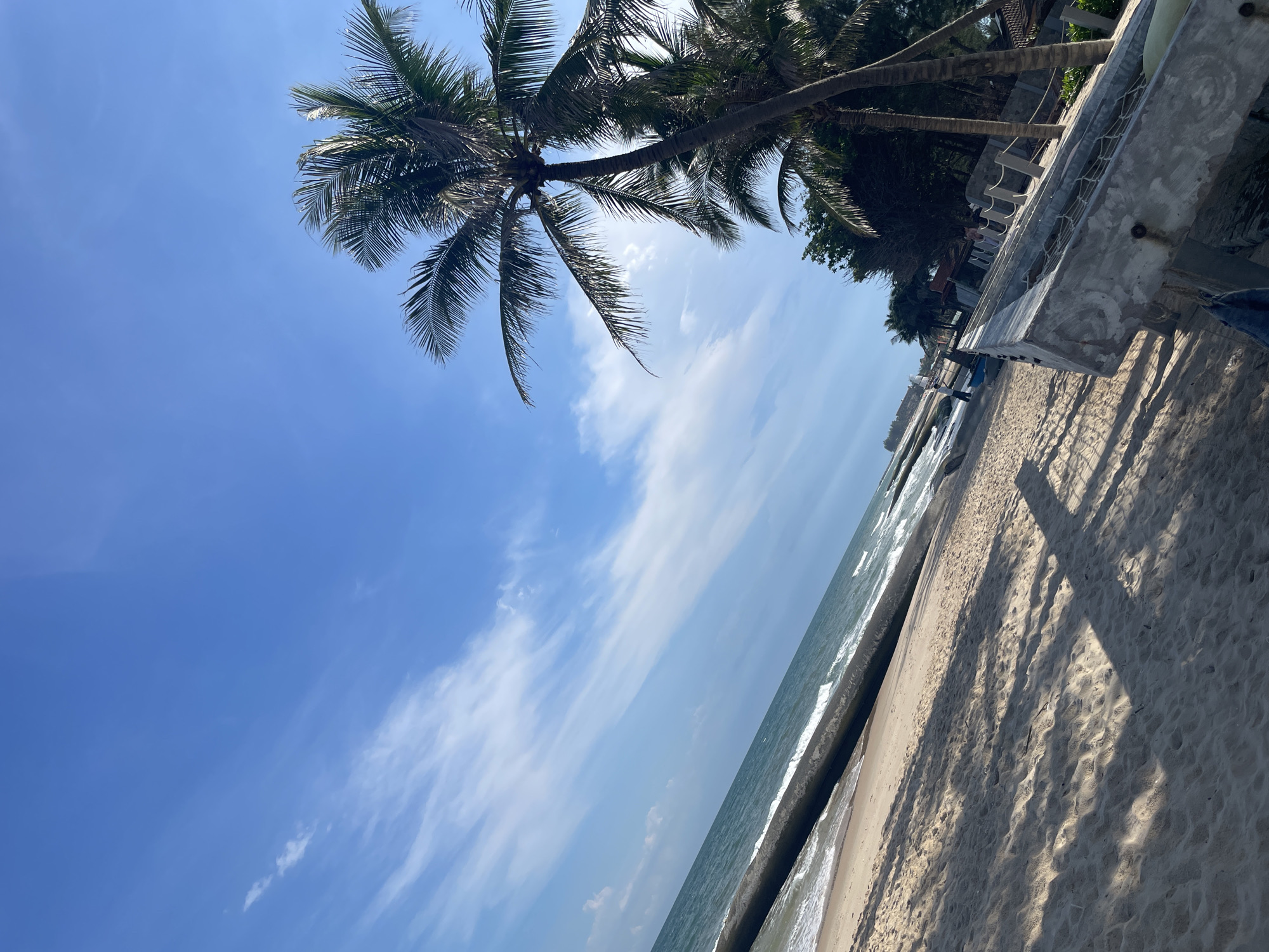 This is a daytime, outdoor, eye-level, full-shot photograph taken at Mui Ne Beach in Phan Thiết, Vietnam. The composition is a diagonal shot looking towards the ocean. The foreground features a sandy beach with distinct shadows cast by an unseen structure. Adjacent to the beach is a low concrete wall with rope railings and what appears to be netting. Beyond the wall, several palm trees stand tall, their fronds silhouetted against a bright blue sky with scattered white, wispy clouds. In the midground, the ocean meets the shore with gentle waves. A dark, linear structure, possibly a breakwater or pier, runs along the water's edge. In the distance, more palm trees and structures are visible, and a person can be seen walking along the beach. The overall environment is that of a tropical beach on a clear day.