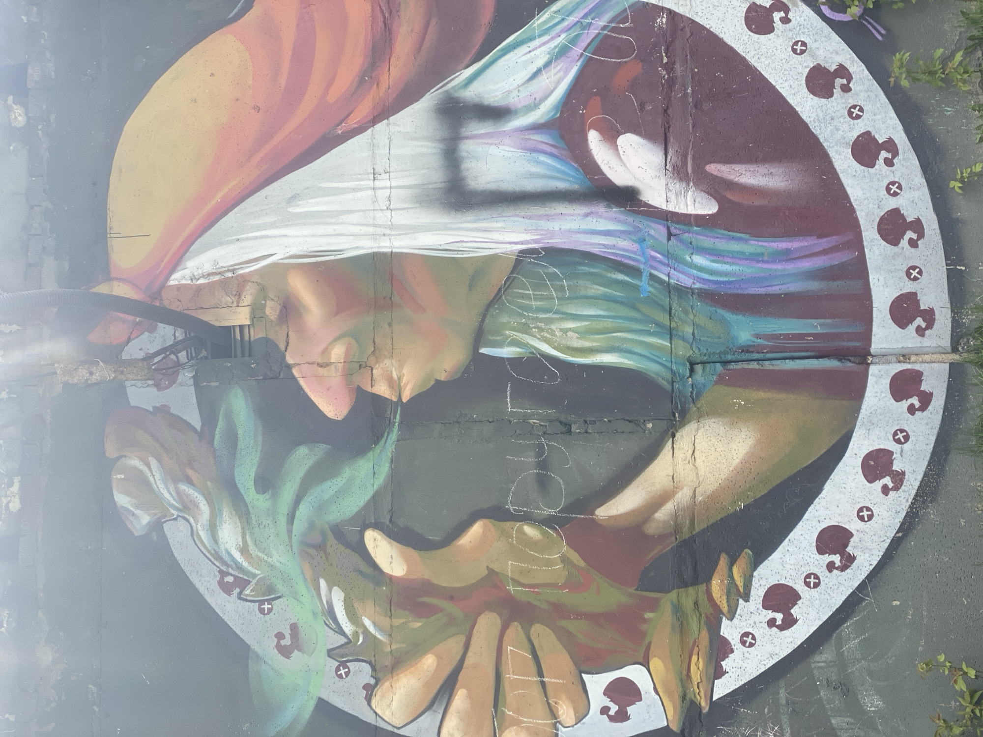 A mural is painted on a concrete wall. It depicts an individual with peach-colored skin and light-colored hair, wearing a white garment. The individual's face is angled downward. Below the face, a pair of hands with peach-colored skin hold a green leafy substance. A circular pattern encircles the mural, featuring mushroom shapes alternating with cross symbols. The concrete wall is gray and weathered. The location of the mural is Novokuznetsk, Russia.