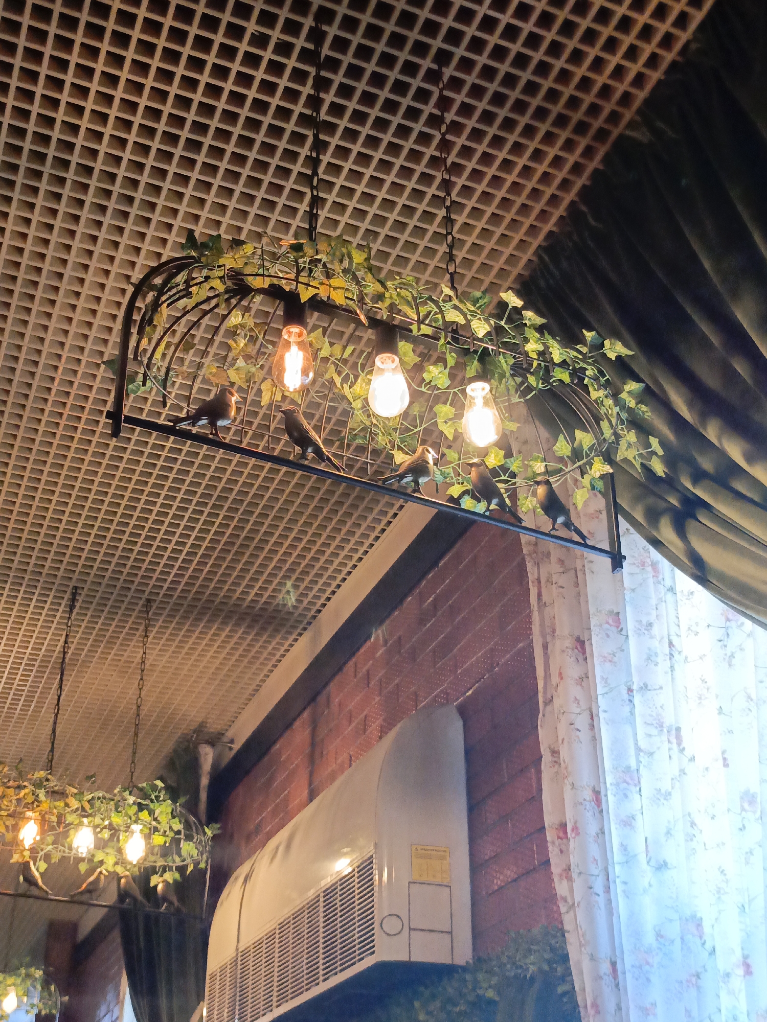 The photograph shows an interior scene with a decorative birdcage chandelier hanging from a square-patterned ceiling. The chandelier features several artificial birds perched on its metal frame, which is adorned with artificial foliage and illuminated by Edison-style light bulbs. Below the chandelier, the image captures a section of a brick wall with an air conditioning unit mounted on it. To the right of the wall, there are floral-patterned curtains. Another similar chandelier can be seen partially in the lower left corner of the photograph. The location is inferred as Verkh-Sosnova, Russia, but this is not directly discernible from the image content.