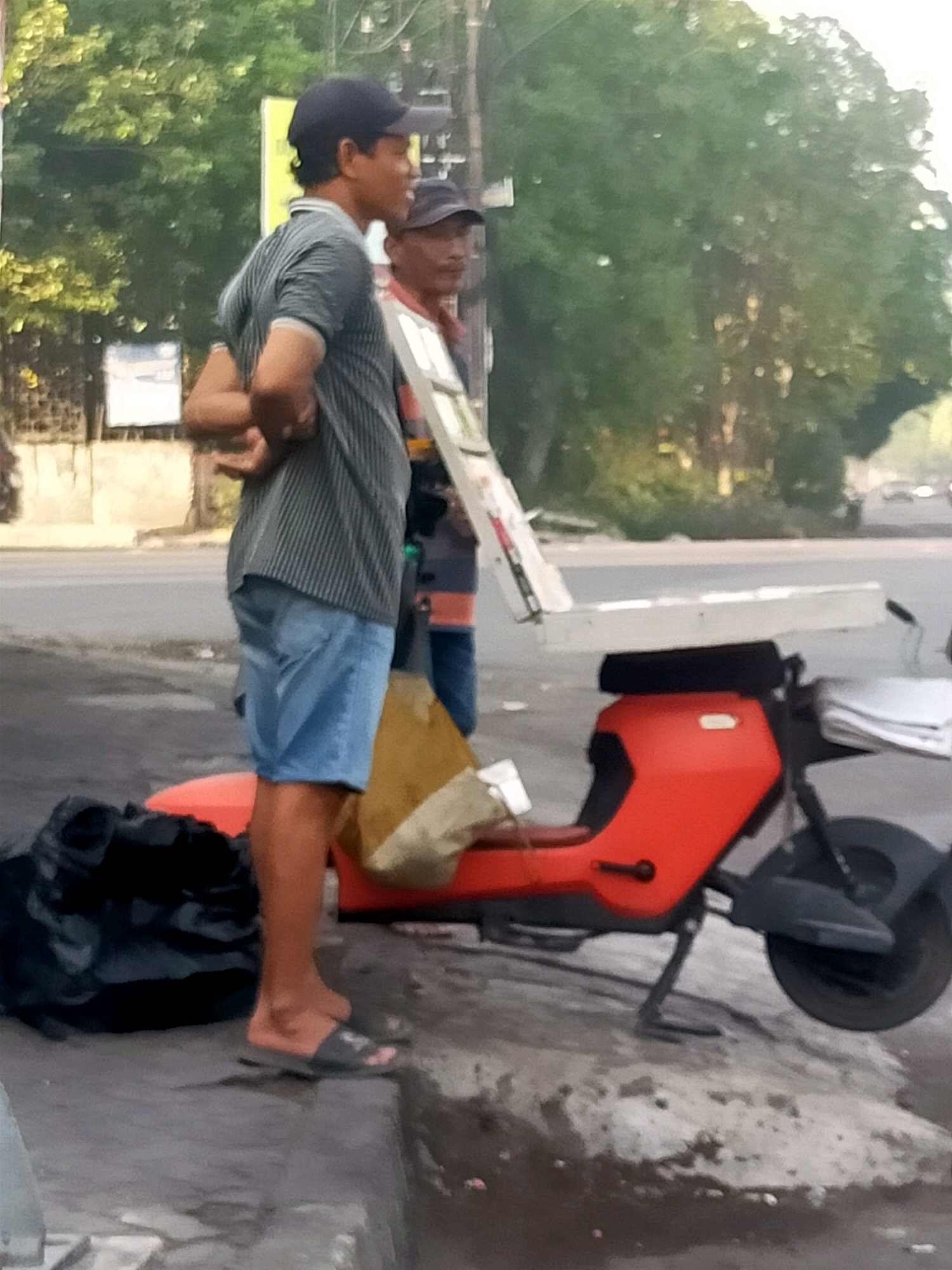 The image depicts two individuals standing on a roadside, likely in Surabaya, Indonesia. The person in the foreground, a male wearing a striped t-shirt and blue shorts, is standing to the left, facing right. He has his arms crossed and appears to be looking away from the camera. To his right, partially obscured, is another male wearing a cap and a striped shirt, holding a white object that resembles a board or a large screen.
In front of the individuals is an orange and black electric scooter. The scooter has a wide, flat white platform extending from the rear, upon which a stack of white papers is placed. A yellow bag is partially visible on the scooter's seat. To the left of the scooter and the foreground individual, a large black bag is on the ground.
The background consists of a road with some traffic, trees, and buildings. The lighting suggests it is daytime, possibly late afternoon due to the warm tones. The overall scene appears to be a street corner or sidewalk area where these individuals are likely engaged in some form of business or activity.