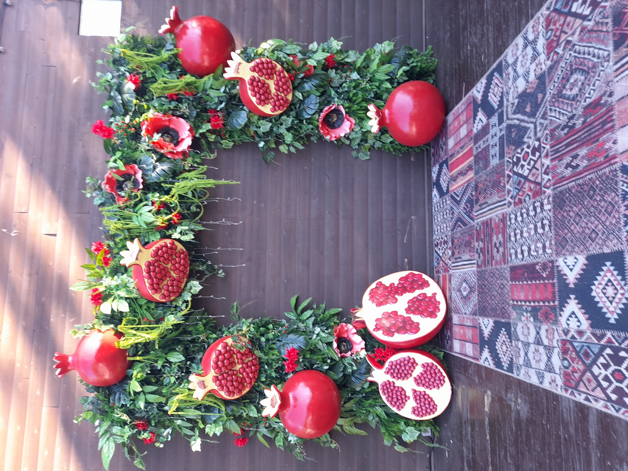 The image depicts a decorative archway adorned with artificial greenery, red pomegranates, and red flowers, set against a brown wooden plank wall. In front of the archway, a carpet with an ethnic pattern is laid out on a wooden floor. The archway is symmetrically arranged with pomegranates and greenery. On the left side, there are two pomegranate halves and a whole pomegranate. On the right side, there are also two pomegranate halves and a whole pomegranate. Red flowers resembling poppies are interspersed with the greenery. The scene appears to be an outdoor decoration, possibly for an event or a storefront. The location is identified as Vladivostok, Russia, although no specific landmarks or context are provided within the image itself to confirm this.