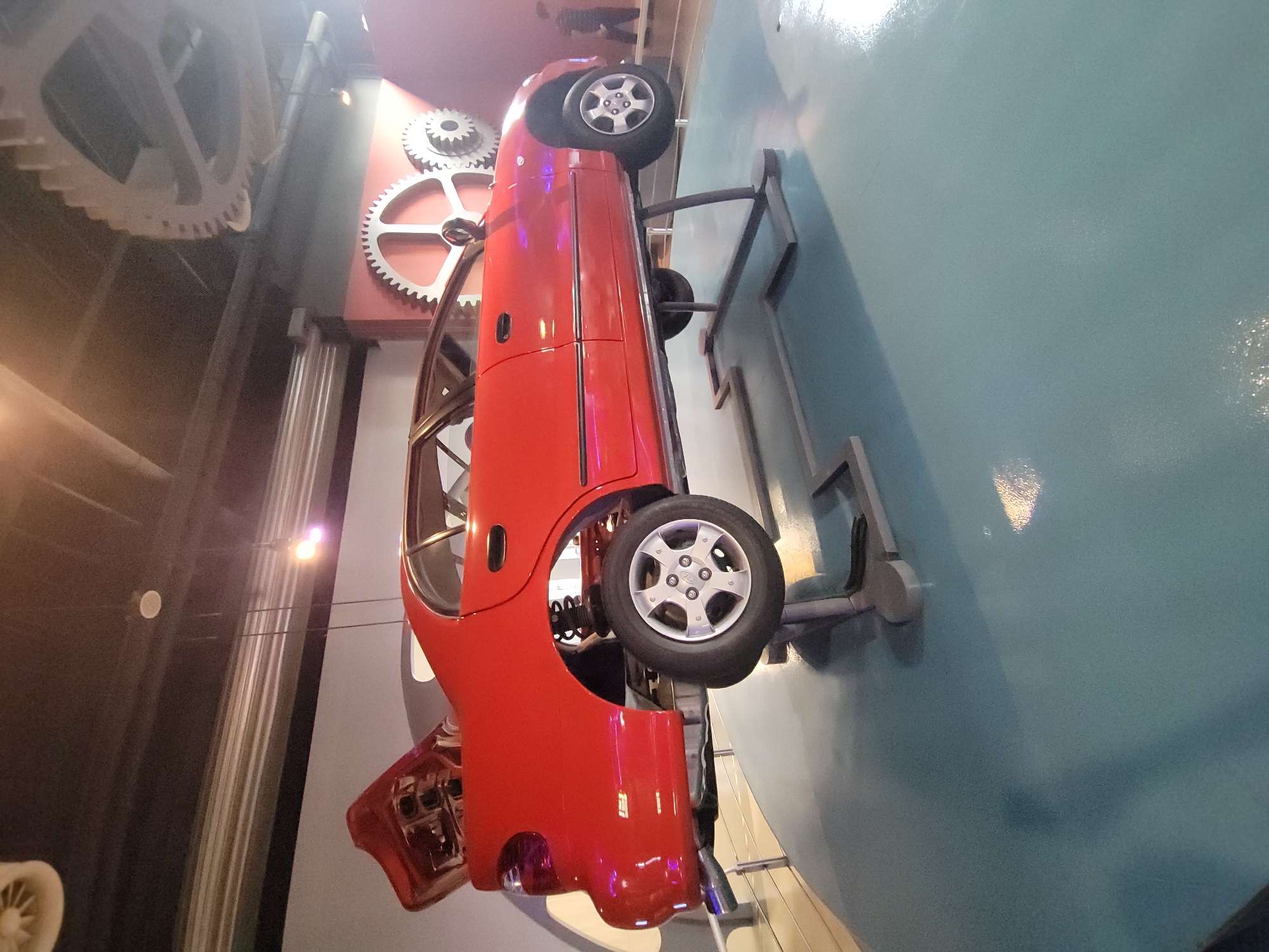 An exhibition display features a red car with its hood and trunk open, elevated on a metal stand above a light blue floor. The car appears to be a sedan, missing some interior components, and its suspension is visible. In the background, large gear-like decorations are mounted on the wall, and several ceiling lights are visible. Two figures, appearing to be staff or visitors, are in the distance. The scene is well-lit and suggests an indoor exhibition or museum setting.