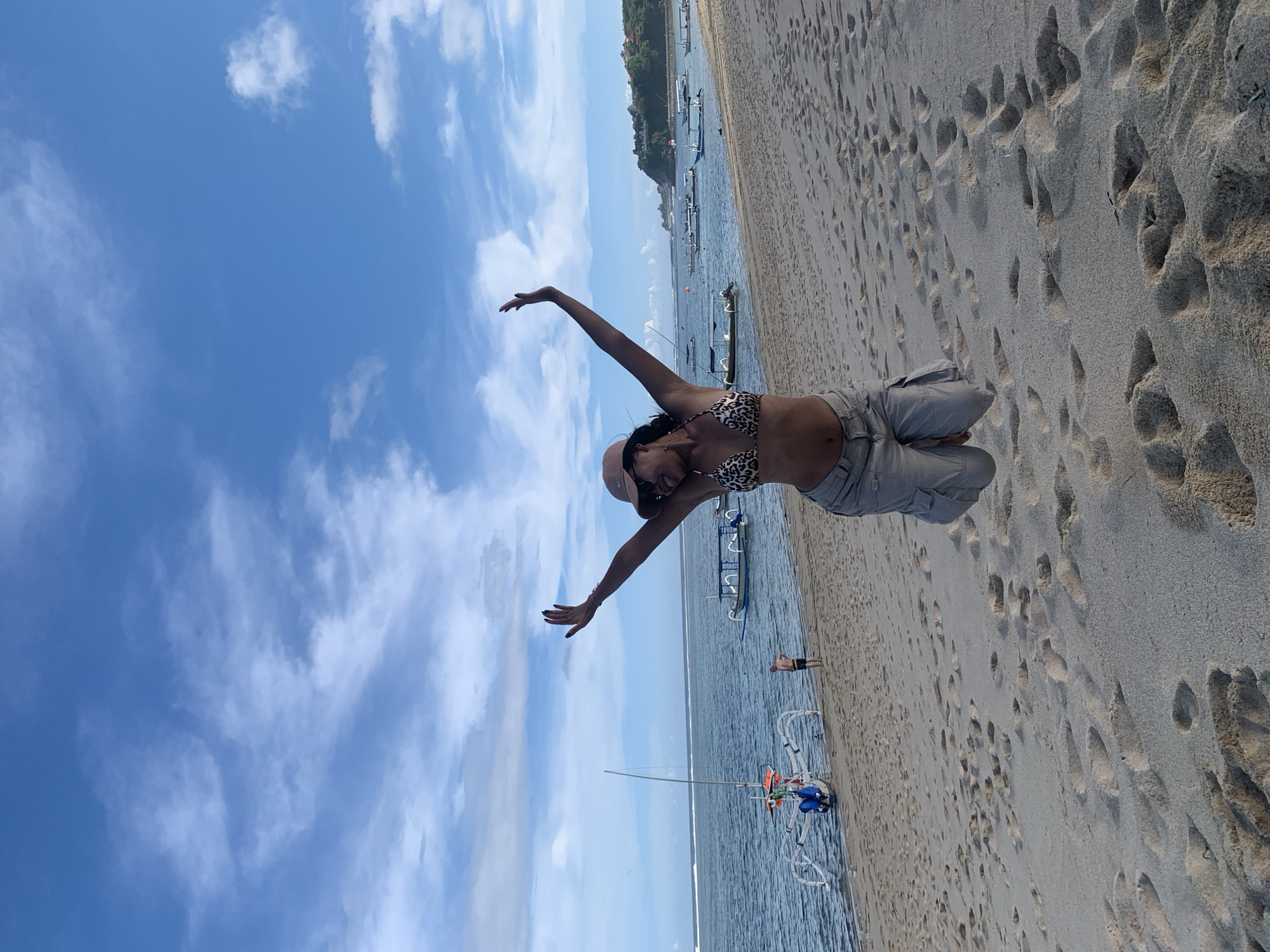 A joyful woman poses on a sandy beach with her arms outstretched, looking upwards and smiling. She wears a leopard print bikini top, light-colored cargo shorts, and a hat. The image captures a bright, sunny day with a clear blue sky dotted with white clouds.
To her right, the calm, blue ocean stretches out, where several traditional-style boats, possibly outrigger canoes, are moored near the shore. One boat features distinctive blue and orange elements. Farther out in the water, a small figure of a person is visible. The beach itself is wide and sandy, covered with numerous footprints, suggesting activity.
The scene depicts a leisurely day at a tropical beach, likely in Kuta Selatan, Indonesia, given the context. The woman's pose conveys happiness and freedom, embracing the bright weather and scenic coastal environment. No visible text is present in the image.