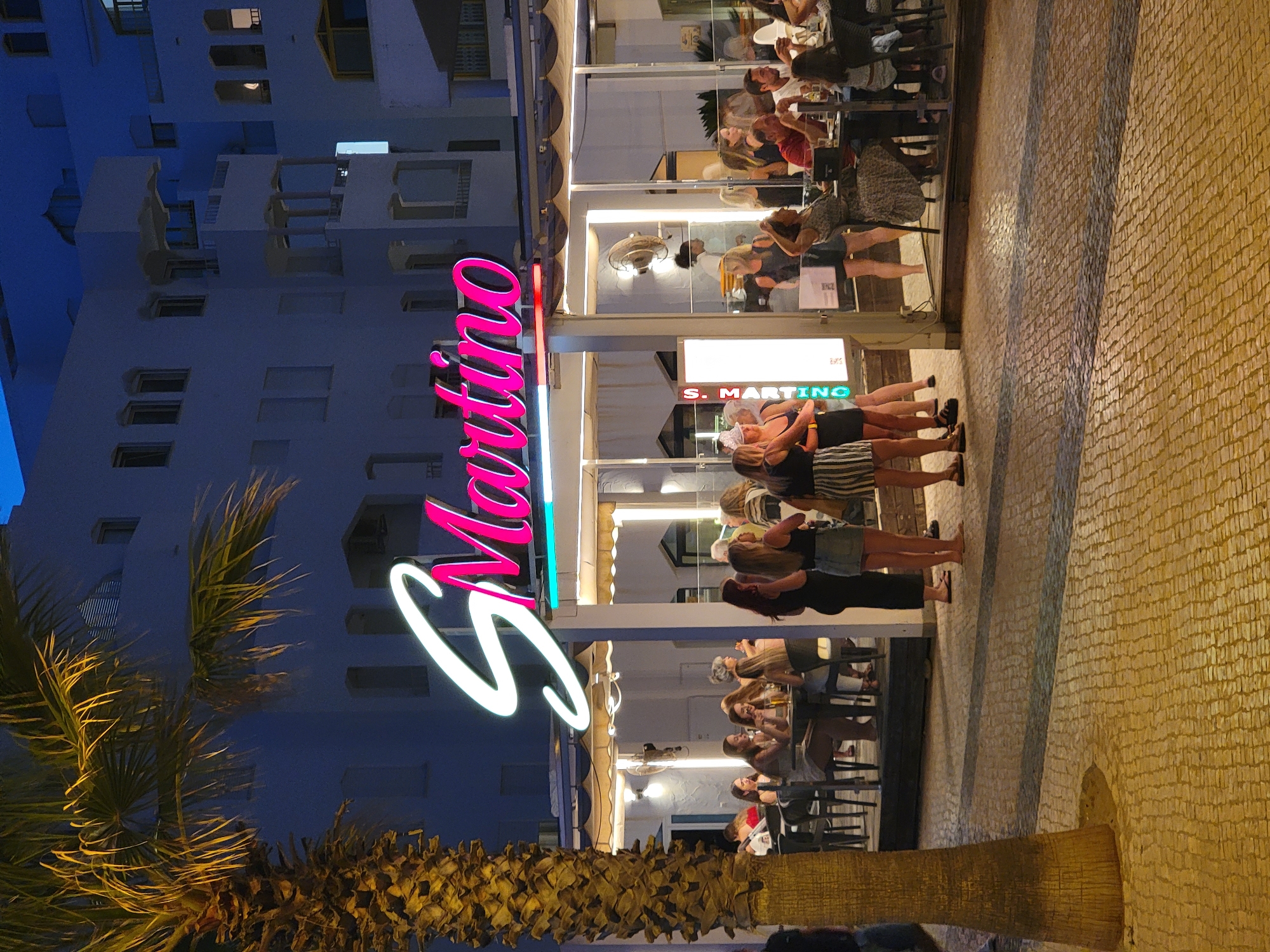 This is a full-frame, eye-level, outdoor, medium shot of a restaurant called "S. Martino" at night in Quarteira, Portugal. The restaurant features a large, illuminated sign with white and pink lettering. Outside the restaurant, tables and chairs are set up on a patterned paved area. Several people are seated at the tables, eating and conversing. Others are standing around the entrance, some appearing to be waiting. A tall palm tree is visible on the left side of the frame, and the building behind the restaurant is a multi-story structure with balconies. The sky is dark, indicating nighttime.