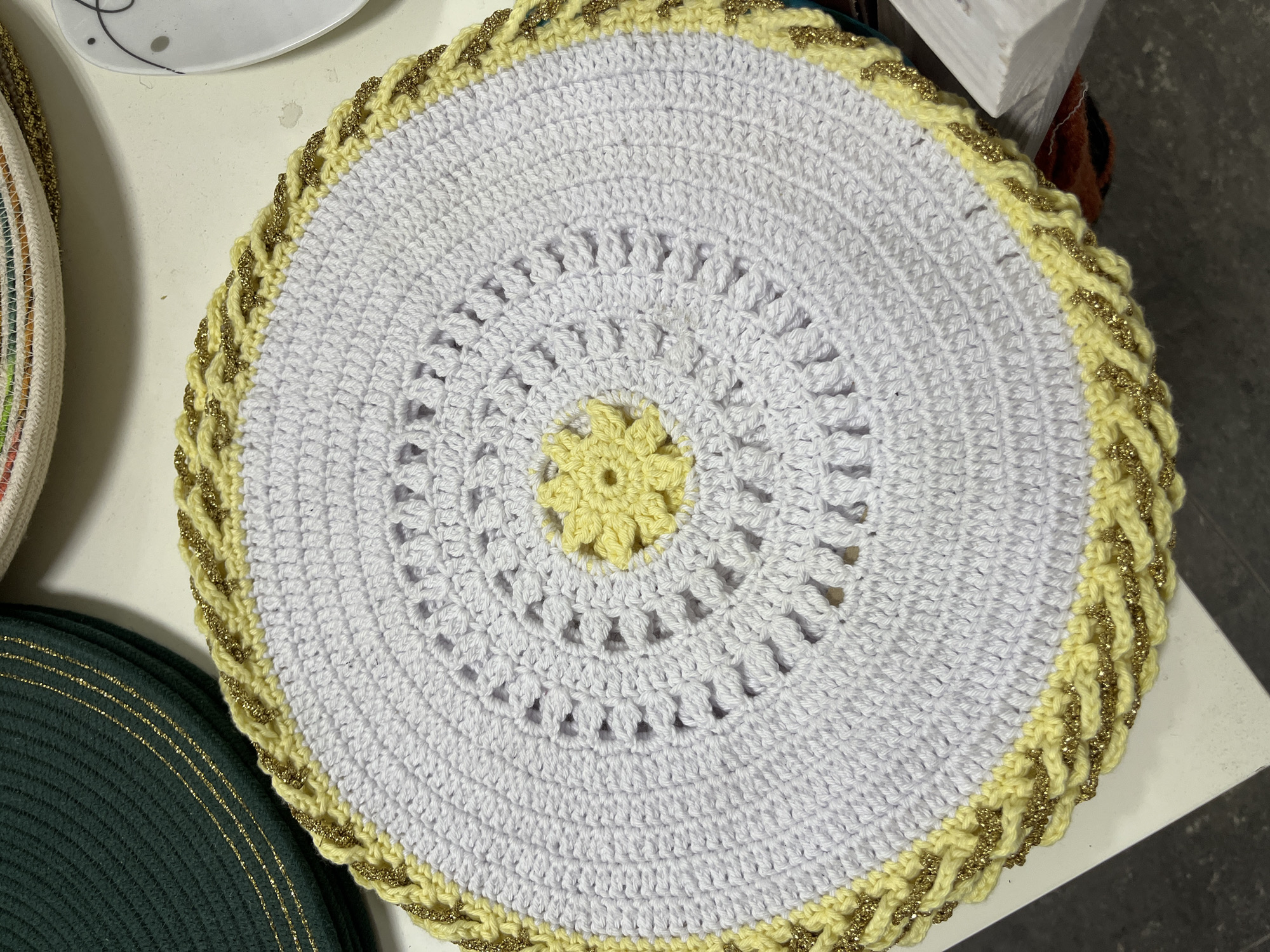 The image shows a close-up, overhead view of a crocheted doily with a white center, a yellow floral design in the middle, and a yellow and gold braided border. The doily is placed on a white surface, likely a table. To the left of the doily, partially visible, are stacked green crocheted placemats with gold trim. To the upper right, a white plate with a black design is visible. The scene appears to be indoors, possibly in a home setting. The specific location within Hrodna, Belarus cannot be determined from the image alone.