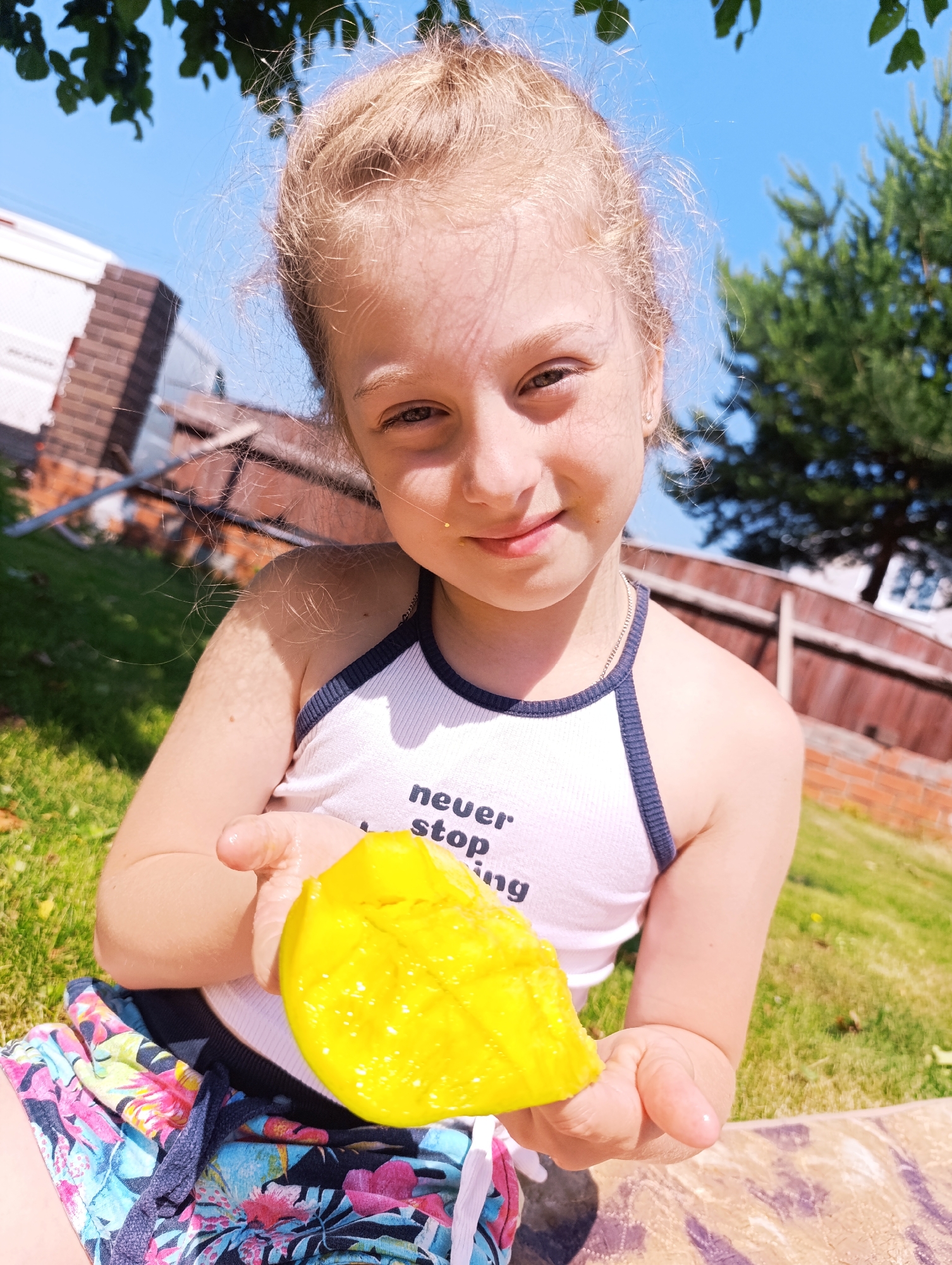 A close-up shot shows a young girl with blonde hair pulled back, holding a bright yellow piece of fruit, possibly mango, in her hands. She is wearing a white tank top with navy trim and a floral-patterned skirt. The girl is outdoors, seated on a patterned blanket, with green grass and trees visible in the background under a clear blue sky. A fence and a brick structure are also present in the background. The sunlight casts shadows on the grass and illuminates the girl and the fruit.