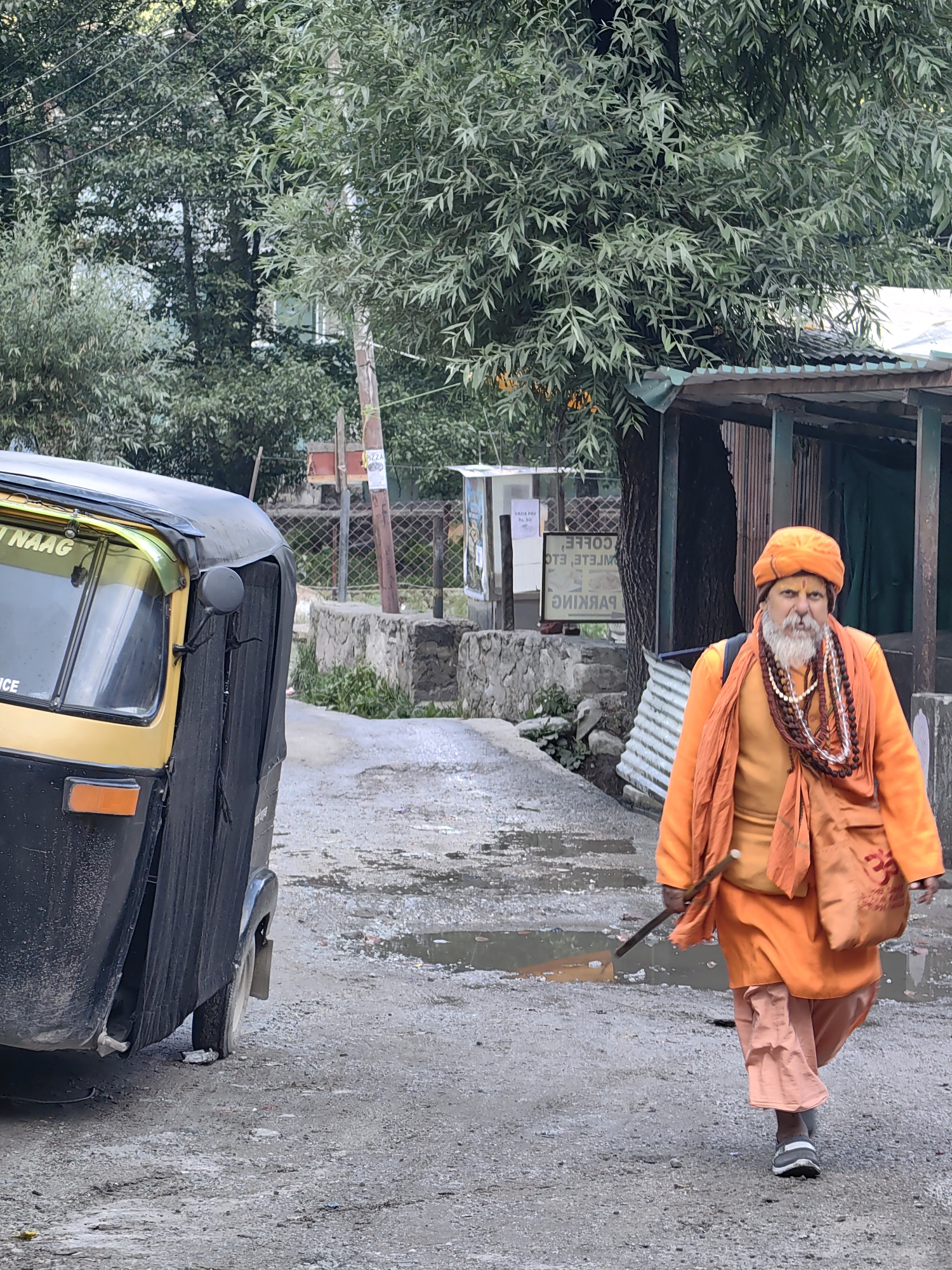An elderly Indian man with a long white beard and a red tilak on his forehead walks down a wet, unpaved street in the city of Goshal, India. He is wearing traditional saffron-colored robes and a matching turban, adorned with prayer beads around his neck. He carries a small bag over his shoulder and holds a stick in his right hand. To his left, a black and yellow auto-rickshaw is parked. The background shows lush green trees and a small, rustic structure with a corrugated metal roof. The street is littered with some debris and has puddles of water, indicating recent rain.