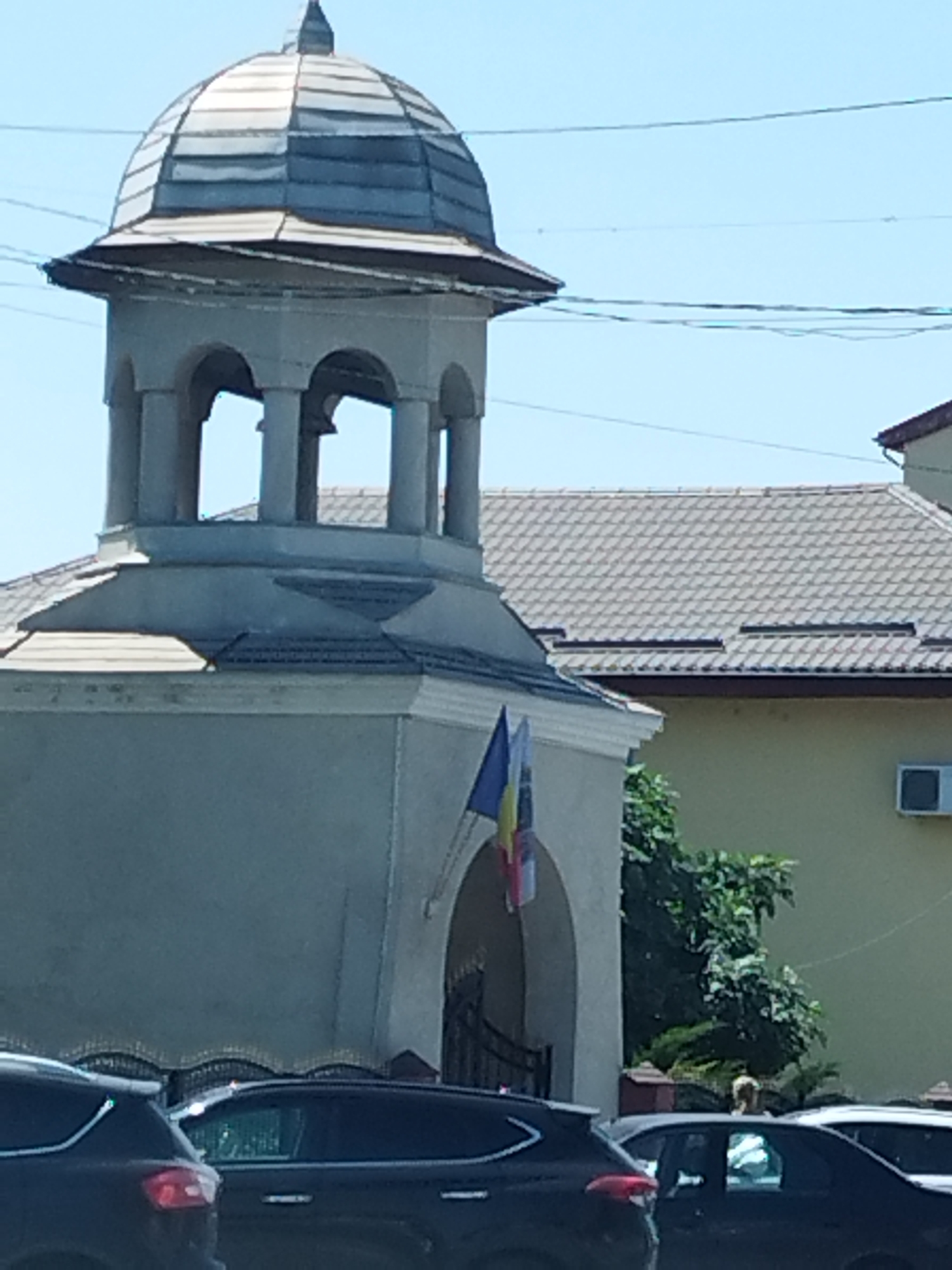 The image shows the exterior of a building with a bell tower. The building appears to be a religious structure, possibly a church, with a dome-shaped roof on the tower and arched openings. Two flags, one blue and yellow and another red, white, and blue, are visible on the facade of the building. A yellow building with a tiled roof is adjacent to the bell tower. Several cars are parked in front of the structures. Trees and foliage are present on the right side of the image, and an air conditioning unit is mounted on the yellow building. The sky is clear and blue. The scene is likely located in Bragadiru, Romania, given the flags and architectural style.
