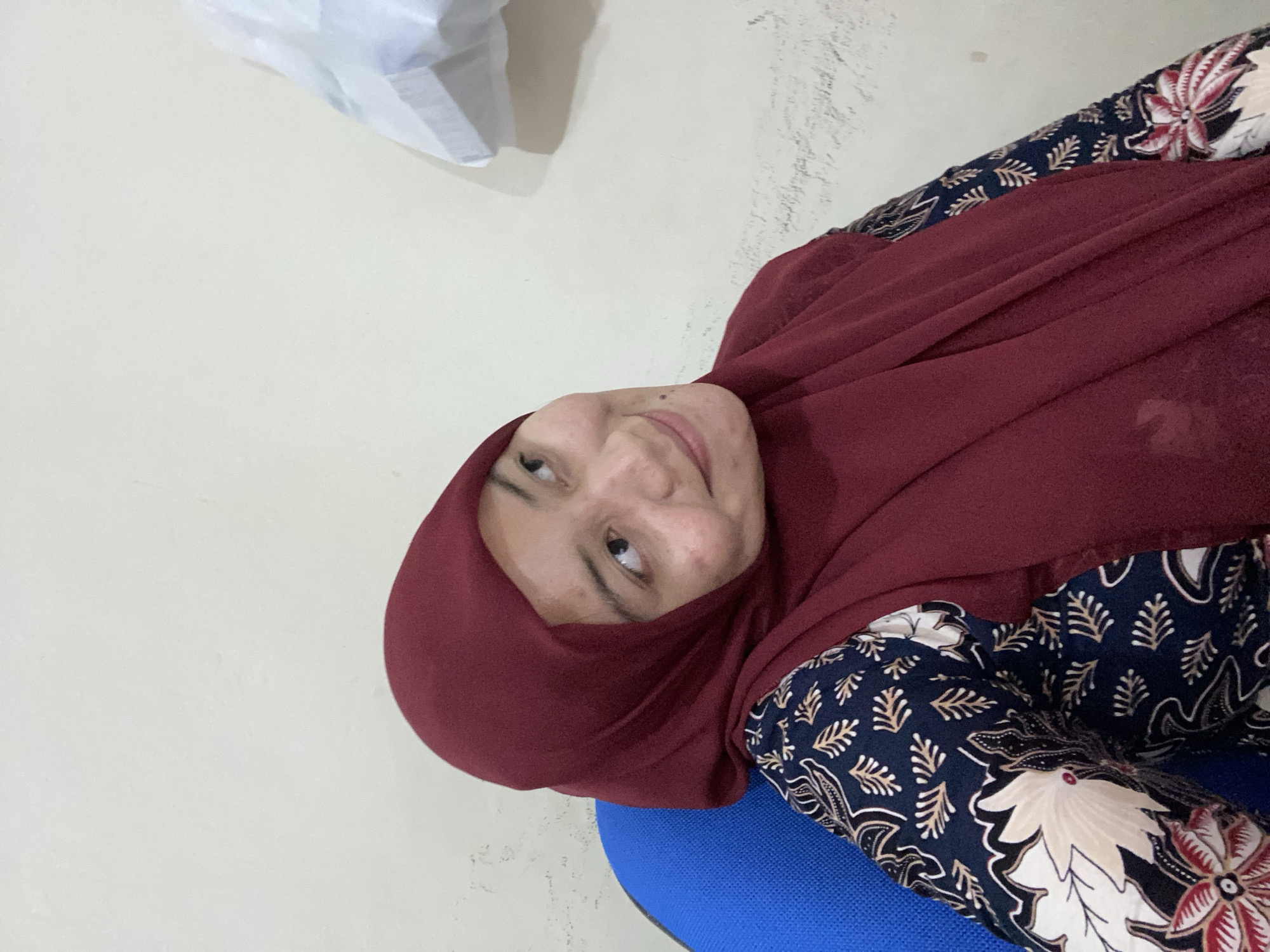 An Indonesian woman wearing a maroon hijab and a dark blue patterned blouse is seated in a blue chair against a light-colored wall. She is looking to her right with a slight smile. A white object is visible on the wall to the upper right of the frame. The overall scene suggests an indoor setting, possibly a home or office, in Banjarbaru, Indonesia.