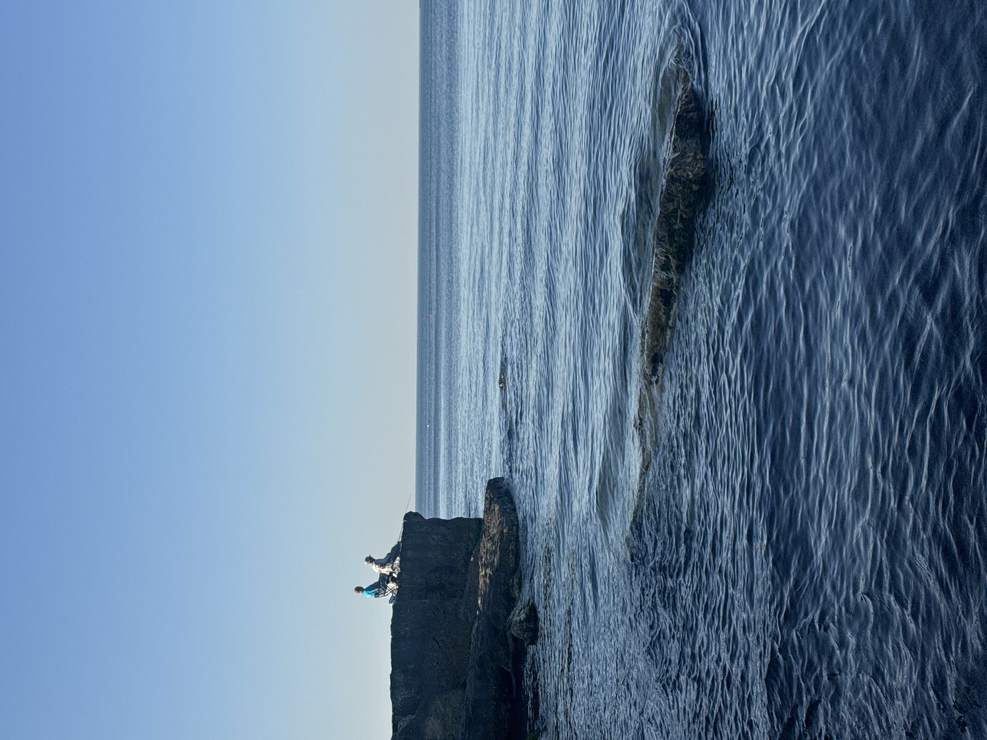 Two individuals are fishing from a rocky outcrop on the shore of the Caspian Sea in Aktau, Kazakhstan. One person is seated in a blue folding chair and is wearing a blue shirt. The other person is also seated and appears to be wearing a hat and a light-colored garment. Both individuals are holding fishing rods, and their lines are cast into the calm, blue water. The rocky formation extends into the sea, with smaller rocks visible in the foreground. The sky is clear and a pale blue, with the horizon distinctly separating the sea from the sky. The overall scene depicts a serene and tranquil moment of recreational fishing by the sea.