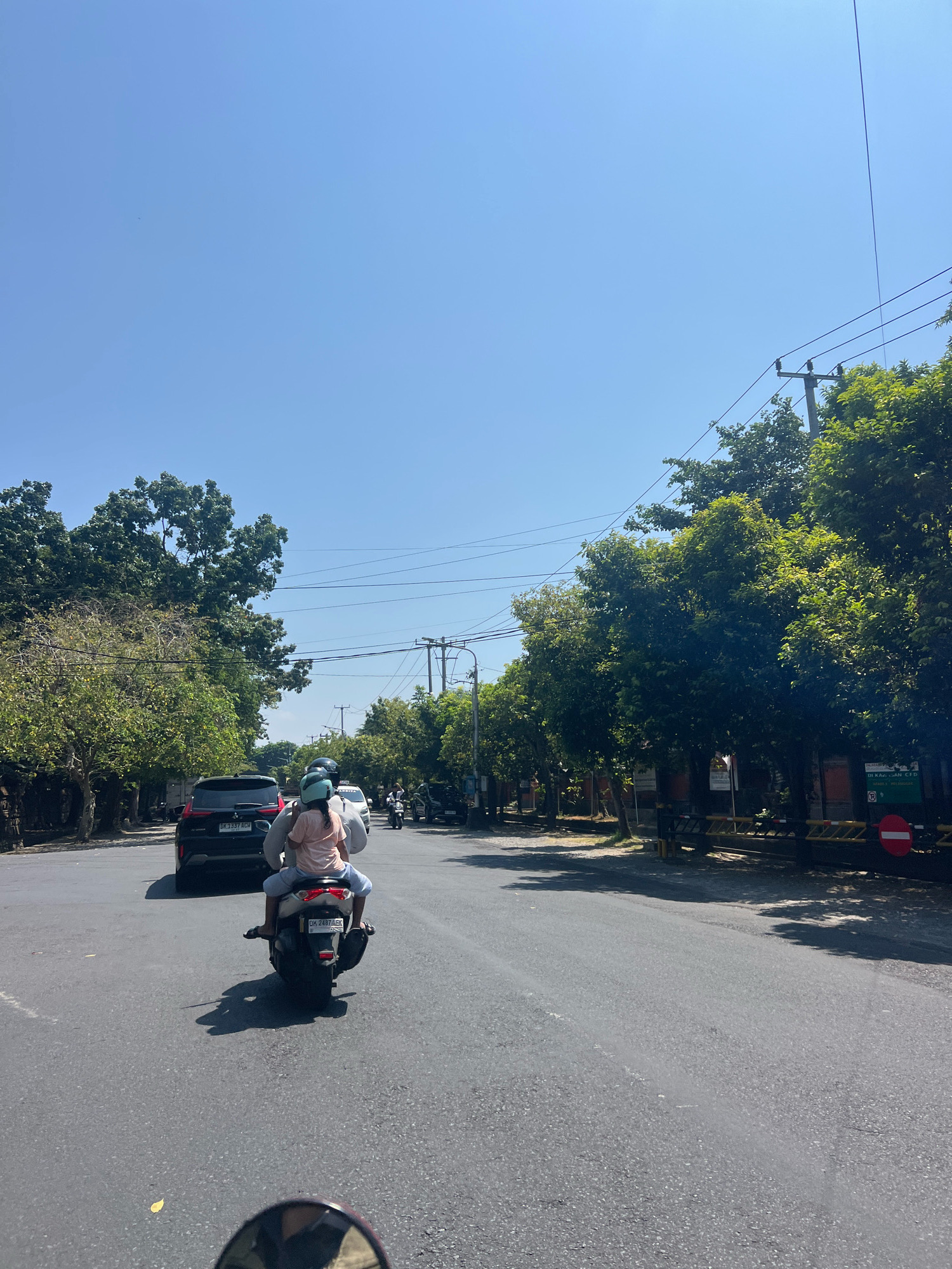 In Denpasar, Indonesia, on a bright, sunny day, a motorbike carrying two individuals, both wearing helmets, travels down a paved road lined with trees. The rider wears a light-colored t-shirt and gray pants, with the passenger behind them wearing a pink t-shirt and light blue pants. A black SUV with license plate "DK 1337 RCH" is ahead of the motorbike. The road has visible lane markings and is bordered by sidewalks and lush green trees. Overhead, power lines stretch across the clear blue sky, supported by utility poles. Signs are visible along the side of the road, including a red and white circular sign with a diagonal line, indicating a prohibition. The scene captures a typical urban streetscape in Denpasar during daytime.