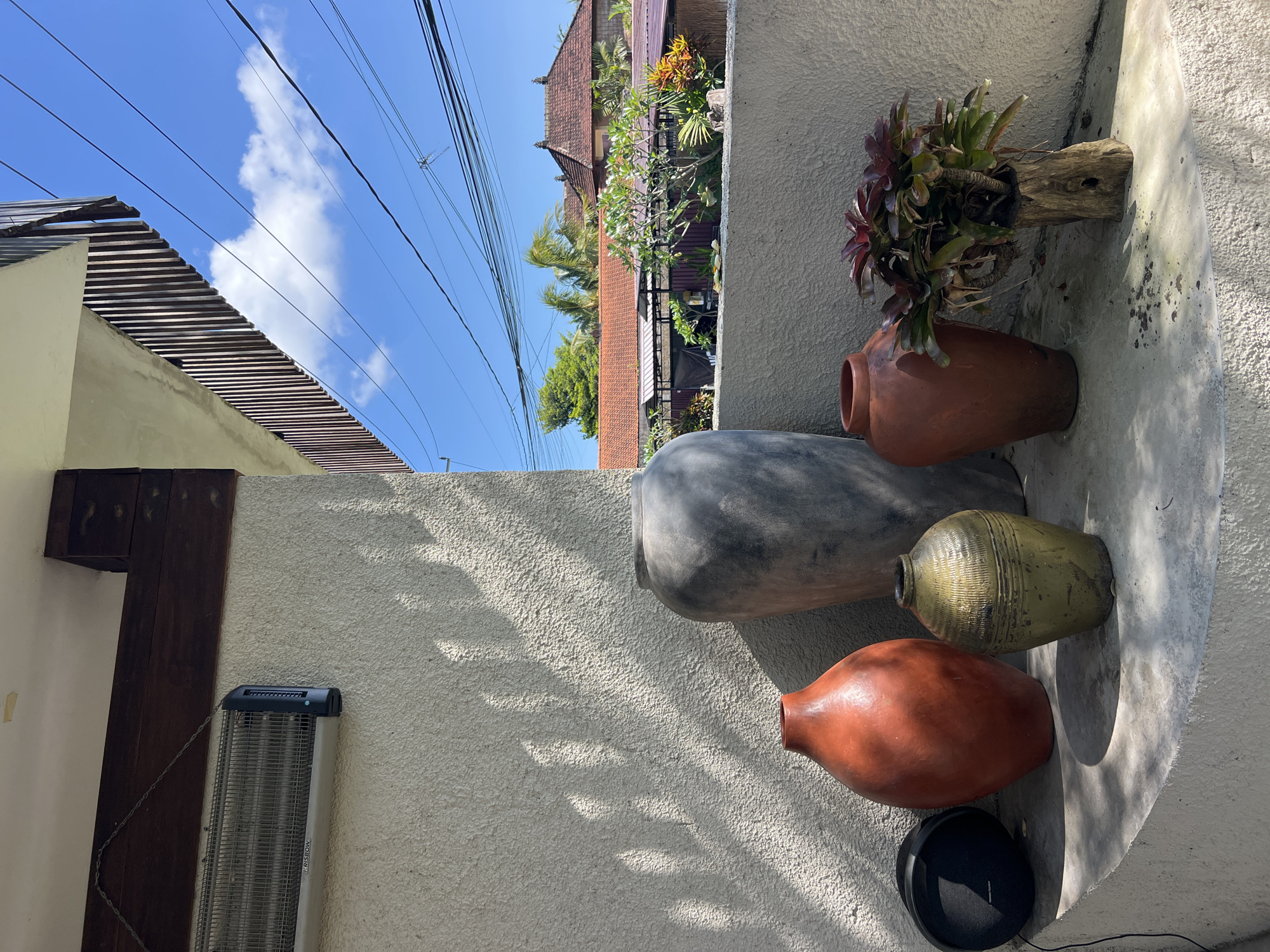 The image depicts a scene in Ubud District, Indonesia, featuring a textured, light-colored wall adorned with various decorative items. Five vases of different shapes, sizes, and colors are arranged on the wall. From top to bottom, these are a reddish-brown vase containing plants, a large gray vase, a smaller olive-green vase, and another reddish-brown vase. A black circular object is positioned at the base of the wall, below the bottommost vase. To the left of the wall, a dark-colored wooden structure is visible, along with a silver-colored, rectangular device that appears to be a heater. Above the wall, a clear blue sky with sparse clouds is visible, along with power lines and rooftops of buildings in the background. The roofs are made of terra cotta tile. The lighting suggests it is daytime, with dappled shadows cast on the wall.