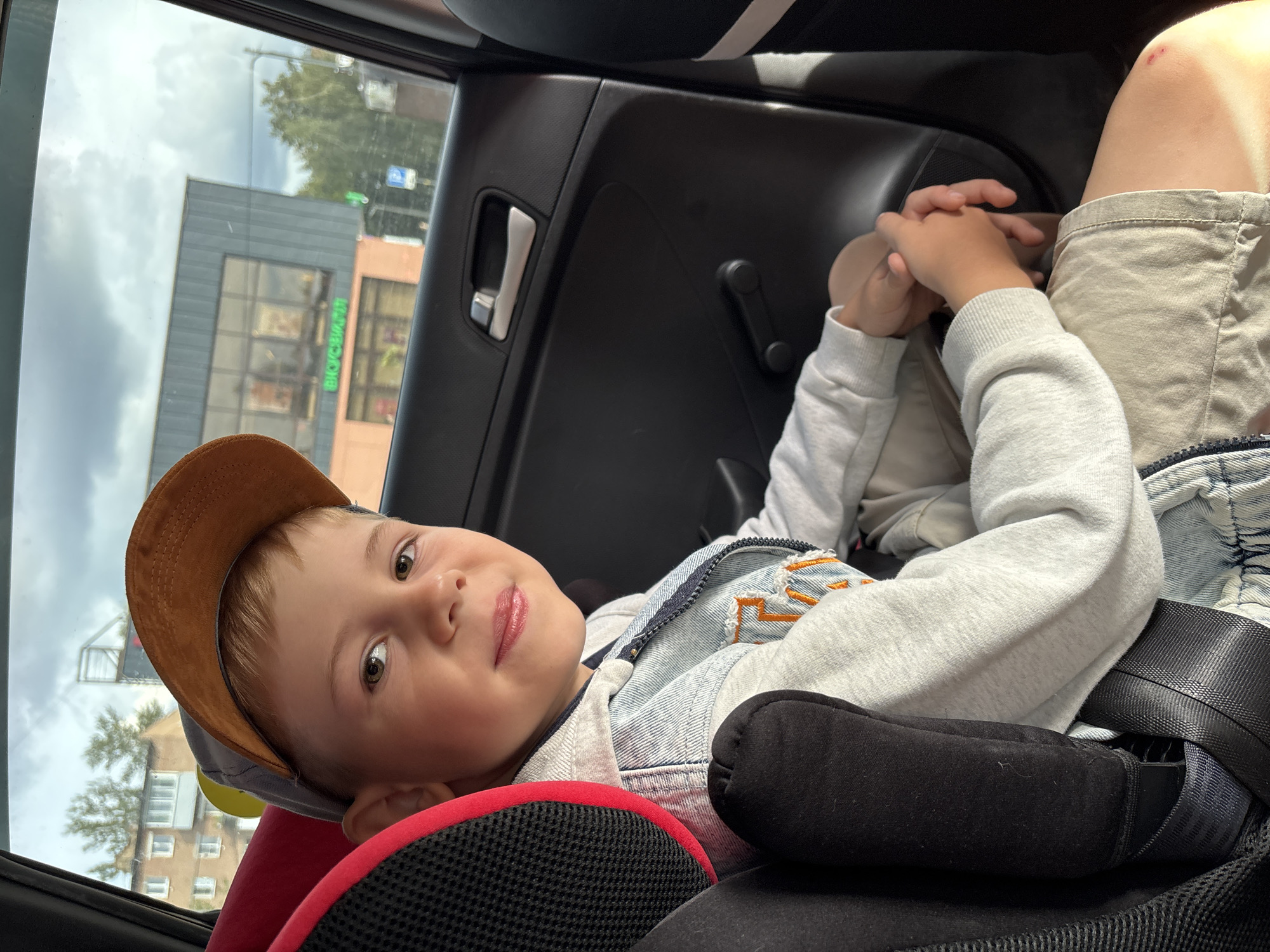 In a car, a young boy with fair skin and light brown hair is seated in a child car seat. He is wearing a light blue denim jacket over a grey hooded sweatshirt, and beige shorts. He has on a brown and grey baseball cap. His hands are clasped in his lap. Outside the car window, a two-story building with a grey facade and large windows can be seen, with green signage reading "BIOCARDI". Trees and a cloudy sky are visible in the background. The car interior shows a black door panel with a silver door handle and a black window crank. The scene is likely in the city of Rusanovo, Russia, as indicated by the presence of the building with Russian signage.
