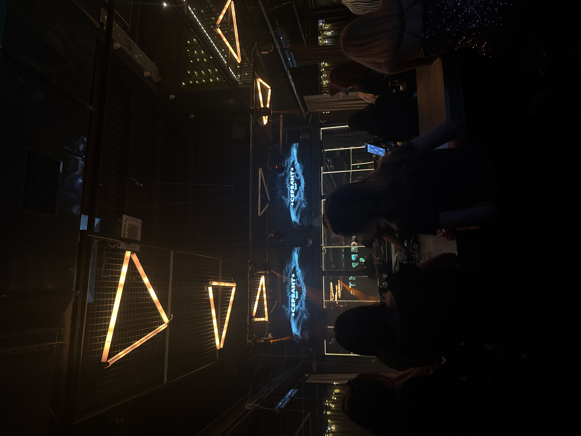 The image captures the interior of "Сервант Бар" (Servant Bar) in Kemerovo, Russia. The scene is dimly lit, featuring an industrial-style ceiling with exposed metal grates and ductwork. Multiple triangular light fixtures, emitting an orange glow from neon-style tubes, are suspended from the ceiling. In the background, two large screens display the "СЕРВАНТ БАР" logo in Cyrillic, set against a swirling blue abstract pattern.
A long bar counter runs across the mid-ground, where multiple individuals are present. Most are observed from behind or in profile, appearing as dark silhouettes against the ambient lighting. There are predominantly female figures, with one person on the far right wearing a garment with visible sparkles. A tablet device is visible on the bar counter. In the deeper background, behind the bar, a male figure is partially discernible. The environment includes additional decorative elements such as string lights on a textured wall on the far right. Ceiling vents and small security cameras are also visible on the industrial ceiling structure.