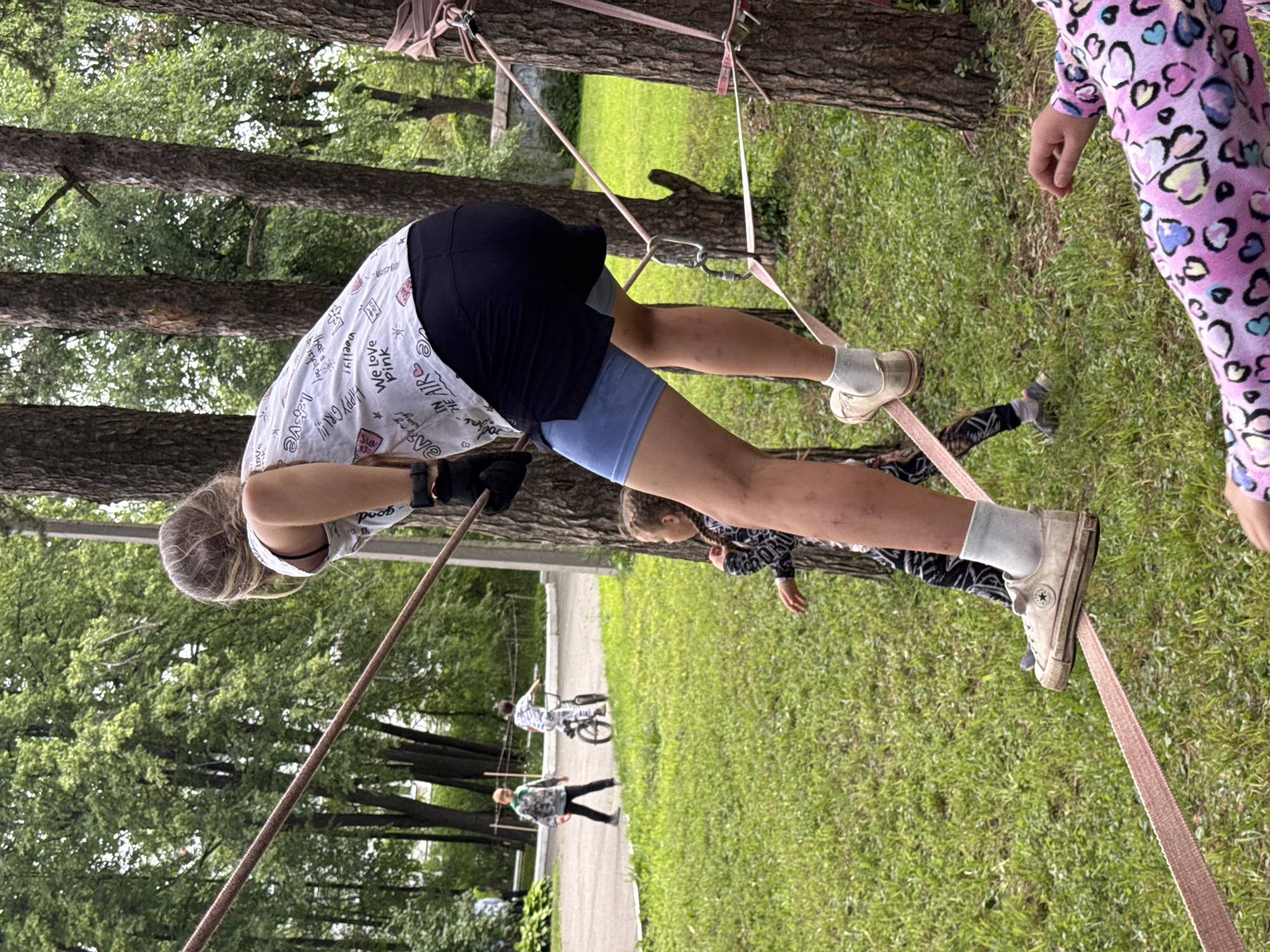 In an outdoor park setting within the city of Chaikovsky, Russia, a group of children are engaged in recreational activities. A teenage girl with blonde hair tied in a ponytail is the central figure, balancing on a slackline suspended between two trees. She is wearing a white t-shirt with a graphic print, black shorts, blue athletic shorts, white socks, and white sneakers. Her arms are bent, and she appears to be focused on maintaining her balance. To her left, a younger girl with braided hair is standing on the grass, looking towards the slackline. Further in the background, two boys are visible on a paved path. One boy is standing, wearing a grey and green jacket and black pants, while another boy is riding a bicycle. The environment is characterized by lush green grass, numerous trees, and a paved pathway. The slackline setup includes ropes and metal carabiners attached to the trees. Another child in the foreground is partially visible, wearing a pink shirt with a heart pattern. The lighting suggests it is daytime, likely overcast, given the soft shadows.