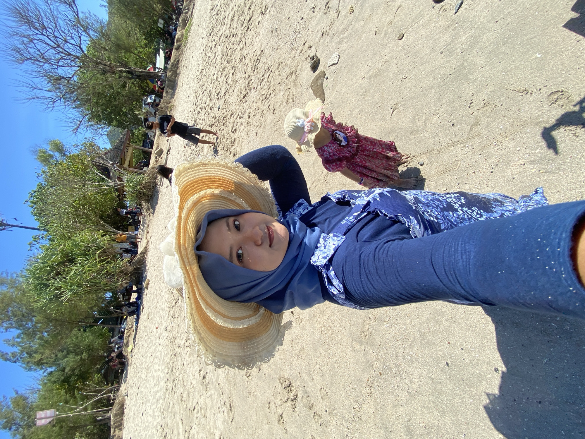 The image shows a woman lying on a sandy beach, wearing a blue hijab, a blue floral patterned shirt, and a large straw hat with a white ribbon. A small child in a red floral dress and matching straw hat is lying near her. A person is walking in the background. The beach is near trees. The location is in the city of Unknown, Indonesia.