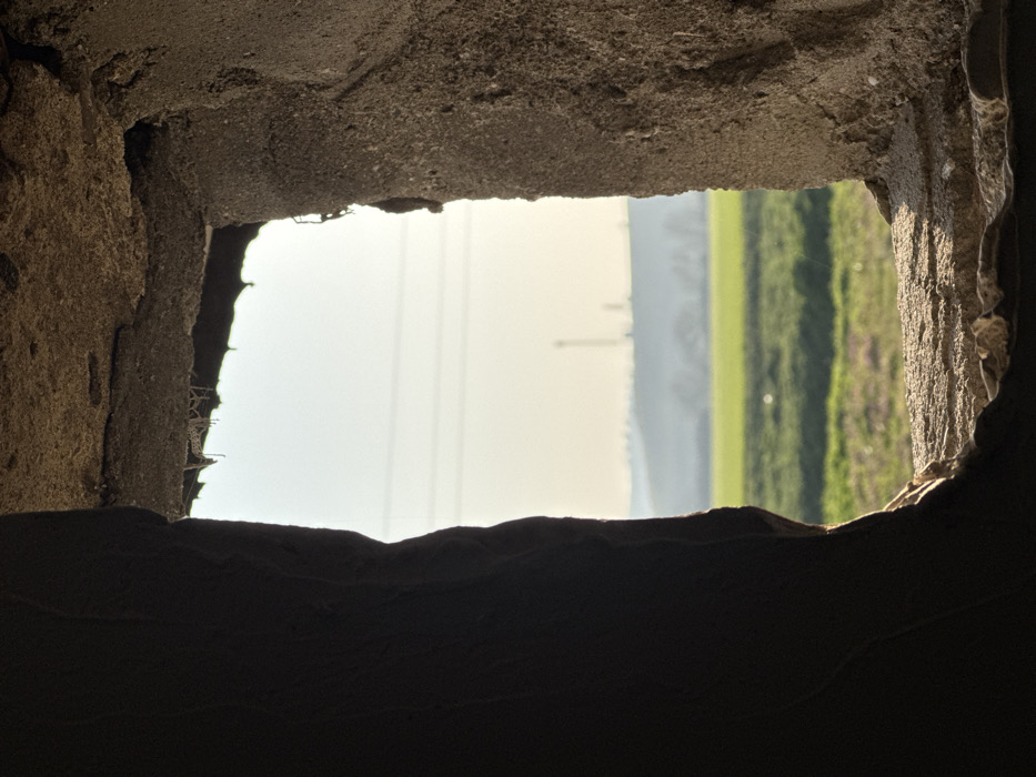 The image displays an exterior rural landscape viewed through an irregular, roughly rectangular opening in a dark, textured wall. The internal surfaces of the opening, likely concrete or stone, are predominantly in deep shadow on the left and top, with partial illumination on the right edge and the bottom sill, highlighting its coarse texture.
Beyond the opening, a brightly lit green agricultural field occupies the foreground and midground. In the distance, a hazy, undulating hill is visible, topped by several vertical structures, including one prominent, tall, slender tower, possibly a communication mast, and smaller industrial or utility structures. Faint horizontal lines, consistent with power lines, traverse the pale, clear sky above the distant hill. No individuals or specific actions are depicted. This scene is located in Rusokastro, Bulgaria.