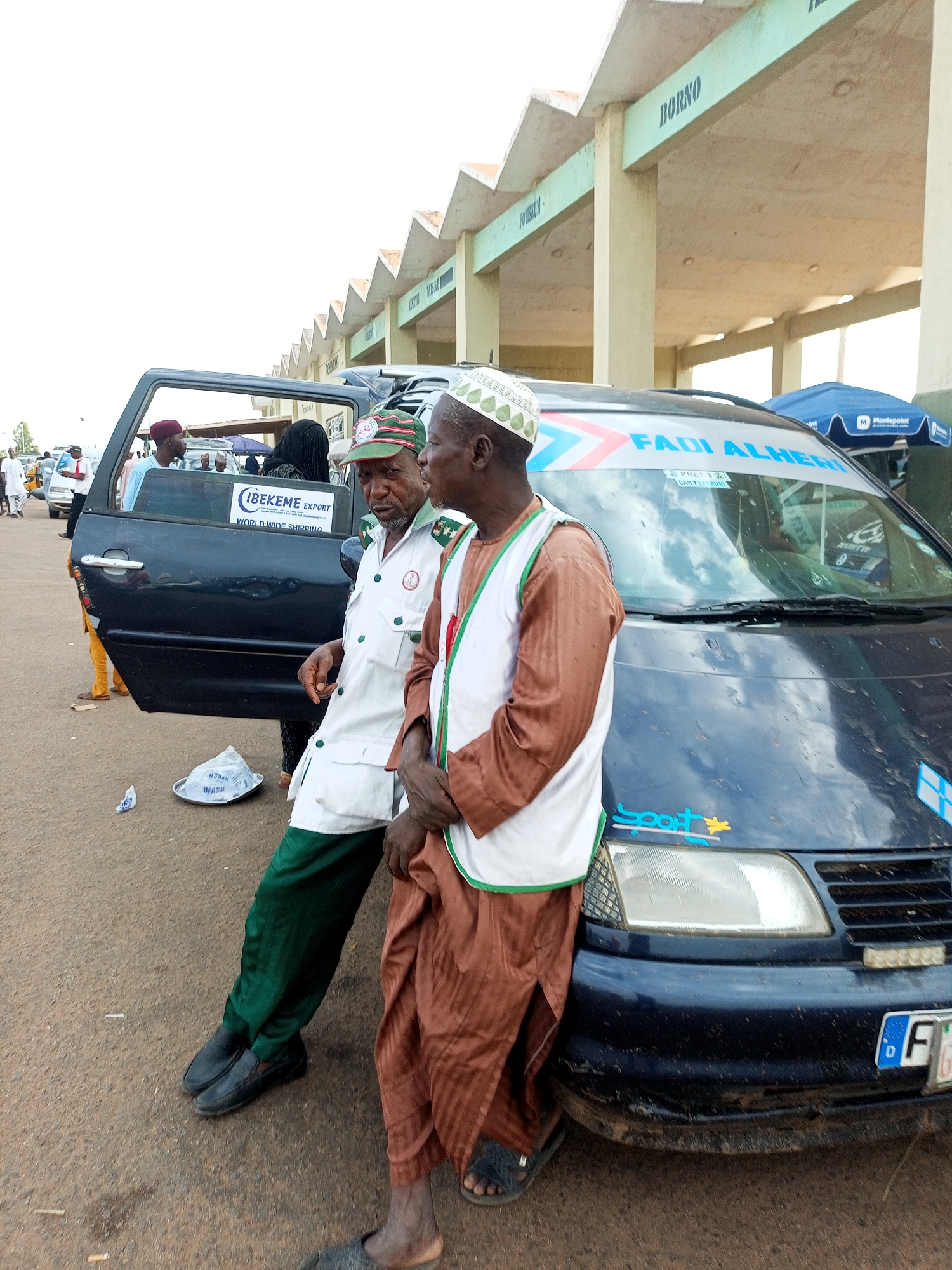 Two men, appearing to be in their 50s or 60s, stand next to a dark-colored van. The man on the left wears a white shirt with green trim, a green cap with a logo, and green trousers. The man on the right wears a brown robe and a white vest with green trim, along with a white cap. They are both looking towards the viewer. The van has the text "FADI ALHERI" in blue and red on its windshield, and a sticker that reads "IBEKEME EXPORT WORLD WIDE SHIPPING". A plate on the front of the van displays "D FG". The scene takes place outdoors, on a paved surface, with a building featuring a repeating triangular roofline visible in the background. This building has text written on it, including "Borno" and "Potskum". The environment suggests a transportation hub or a market area in Gombe, Nigeria.