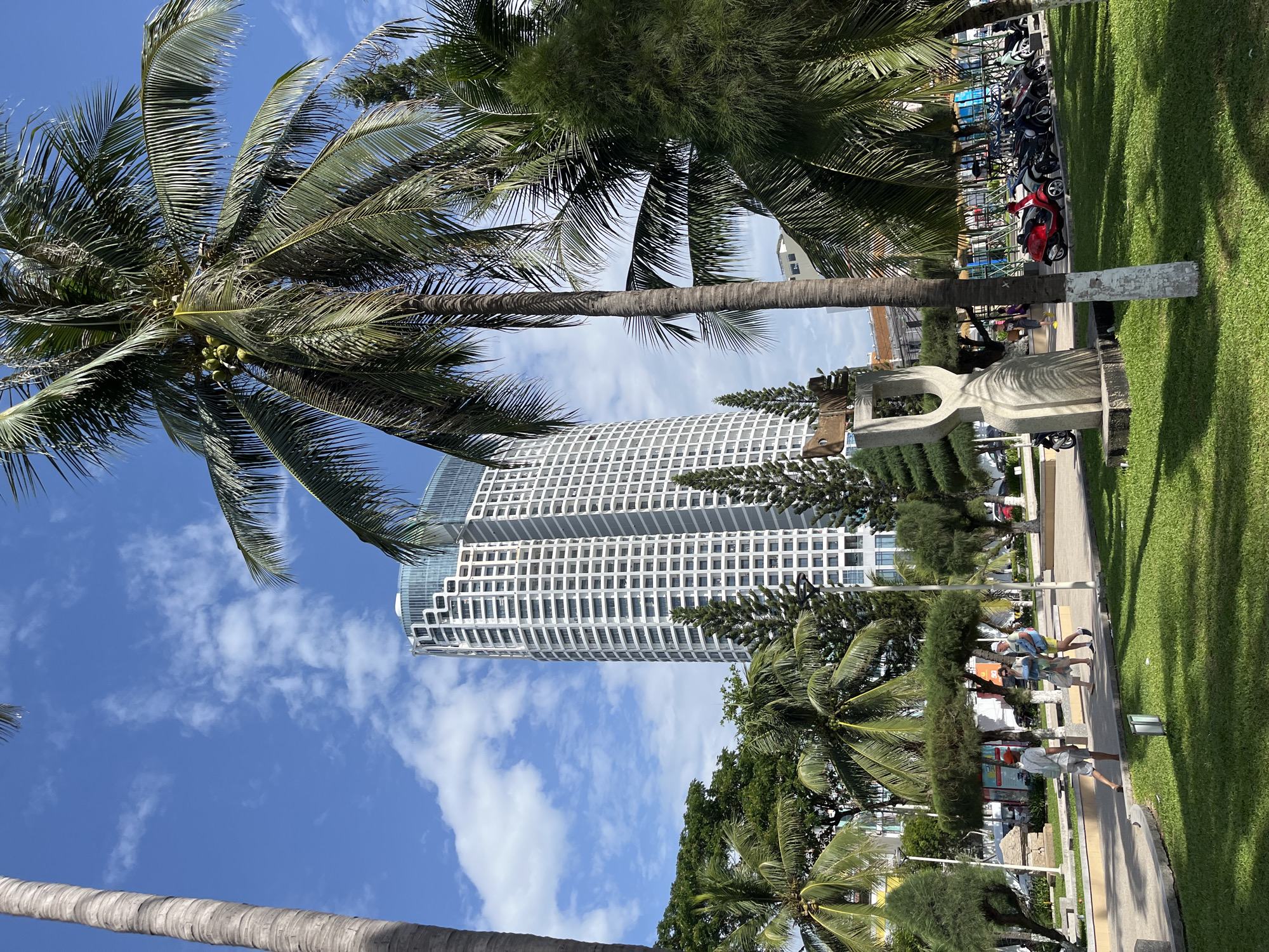 In Nha Trang, Vietnam, a tropical urban landscape unfolds under a partly cloudy sky. A modern, white high-rise building with a distinctive curved facade and blue-tinted windows dominates the background. In the foreground, lush green grass carpets a park area, with several palm trees and other greenery providing shade and a sense of tropical ambiance. A sculptural piece made of light-colored stone stands prominently in the park. People are seen walking through the park, some casually dressed for a warm day. Beyond the park, a row of parked motorcycles, including a bright red one, suggests urban activity. The scene captures a typical day in a coastal Vietnamese city, blending modern architecture with natural elements and everyday life.