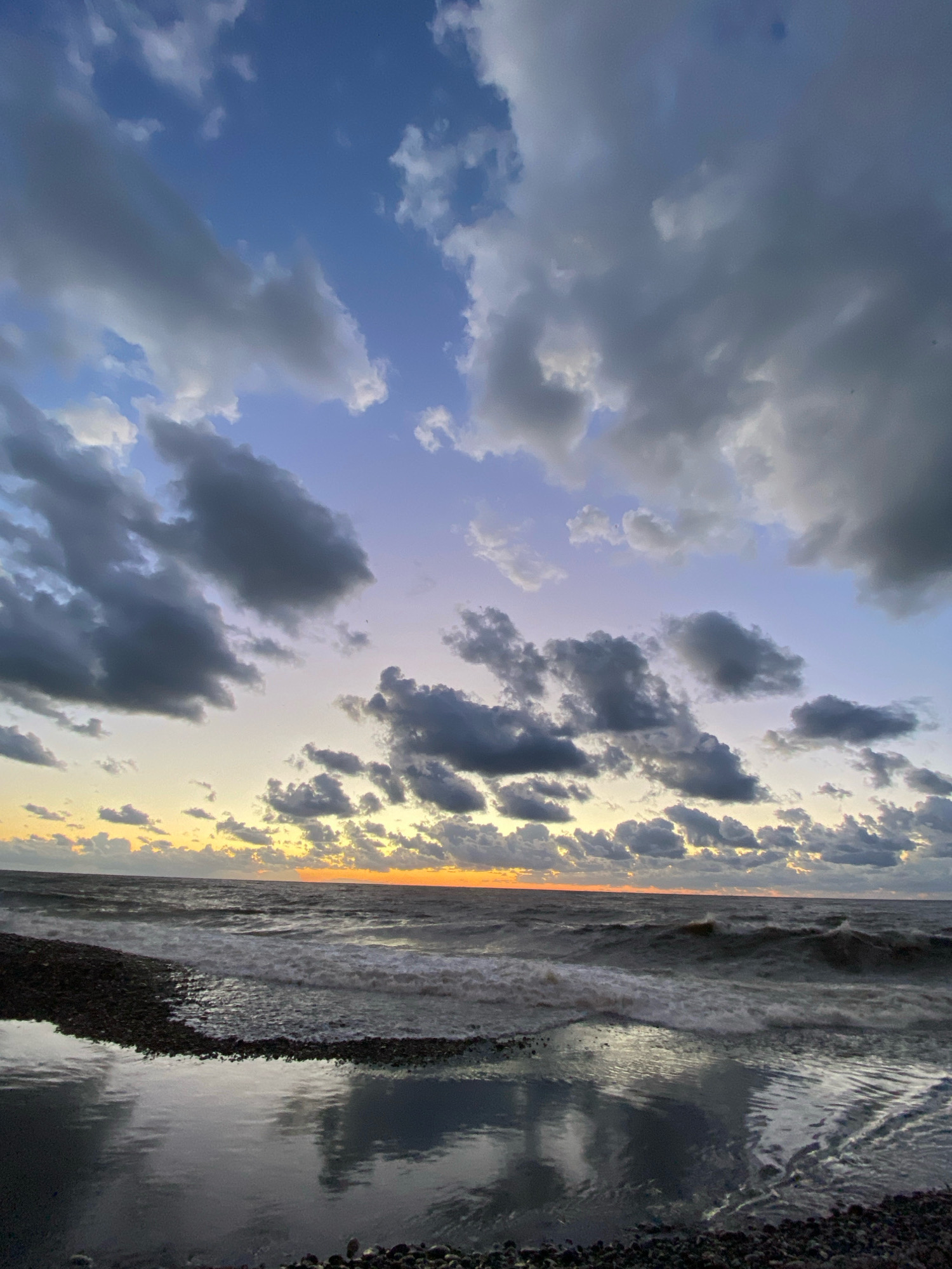 An eye-level shot captures a dramatic sunset over a rocky beach and turbulent ocean. The sky is a canvas of deep blues transitioning to soft purples and pale yellows near the horizon, adorned with scattered, dark grey clouds. The sun, a vibrant orange, peeks through the clouds at the far right, casting a warm glow on the water. Waves crash onto the shore, creating white foam and a shimmering reflection in a calm pool of water on the dark, pebble-strewn beach. The scene is devoid of any people or man-made objects, presenting a raw and natural seascape.