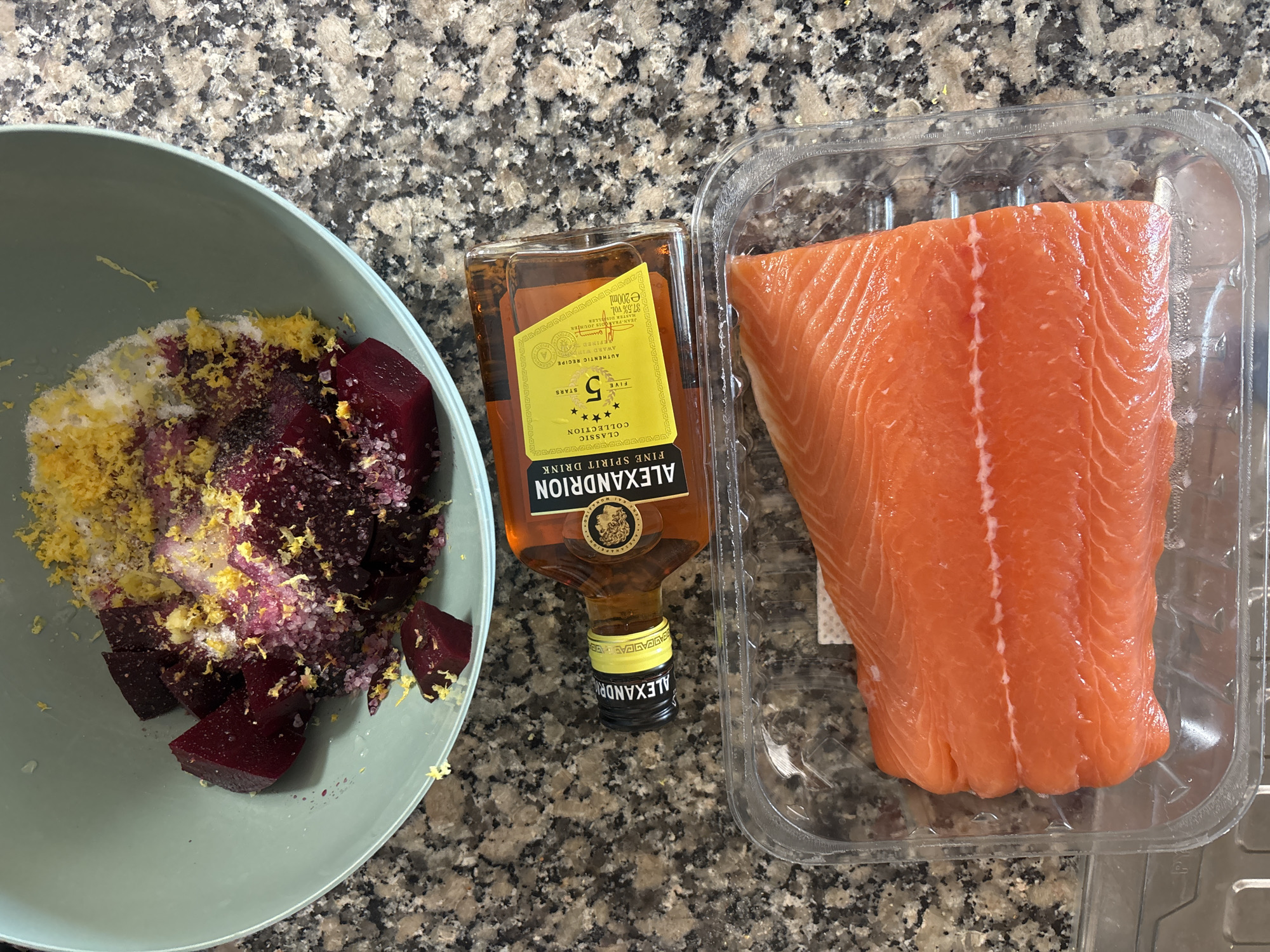 The image shows a top-down view of ingredients for a meal laid out on a granite countertop. In a light green bowl, chopped beets are mixed with salt, pepper, and lemon zest. Next to the bowl is a small glass bottle of Alexandrion 5 Stars Fine Spirit Drink. Below the bottle is a clear plastic container holding a raw fillet of salmon. The scene appears to be indoors. There is no information in the image to suggest a location in Tavira, Portugal.