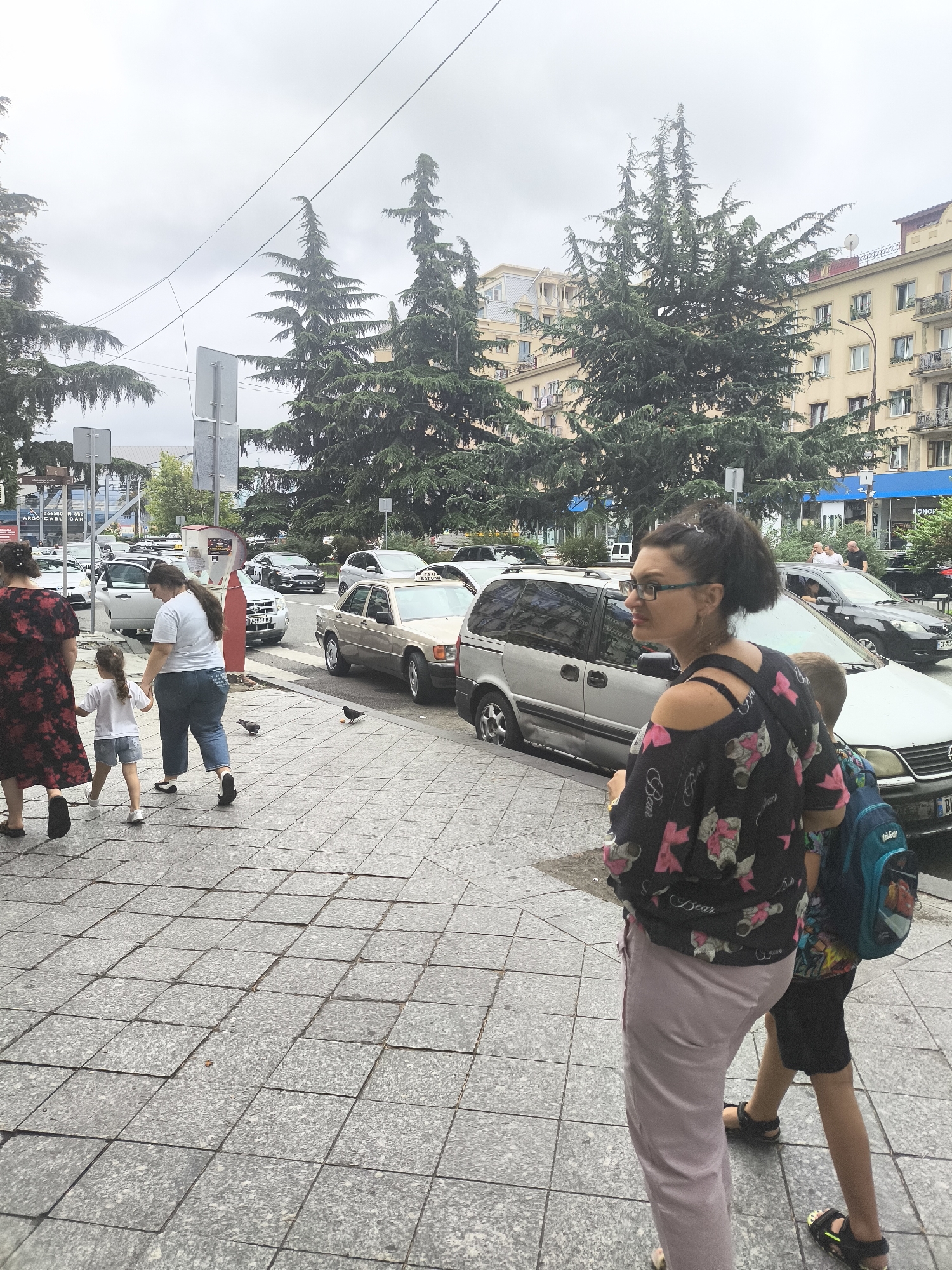 The image captures a street scene in Batumi, Georgia, likely on a cloudy day. In the foreground, a woman with dark hair pulled up in a bun, wearing glasses and a black off-the-shoulder top with a pink floral pattern, walks away from the camera. Beside her, a boy with a blue backpack walks in the same direction, wearing a t-shirt and shorts.
Behind them, further to the left, three people are walking on a tiled sidewalk. A woman in a red floral dress walks with a young girl in a white t-shirt and denim shorts, holding her hand. Another woman in a white t-shirt and blue jeans walks beside them.
The street is lined with parked cars, including a silver minivan and a light-colored sedan. Several pigeons are visible on the sidewalk. In the background, tall evergreen trees are prominent, and apartment buildings with balconies and windows can be seen. Traffic signs and streetlights are also present, suggesting an urban environment. The overall impression is a casual moment in a city street, with pedestrians and vehicles coexisting.