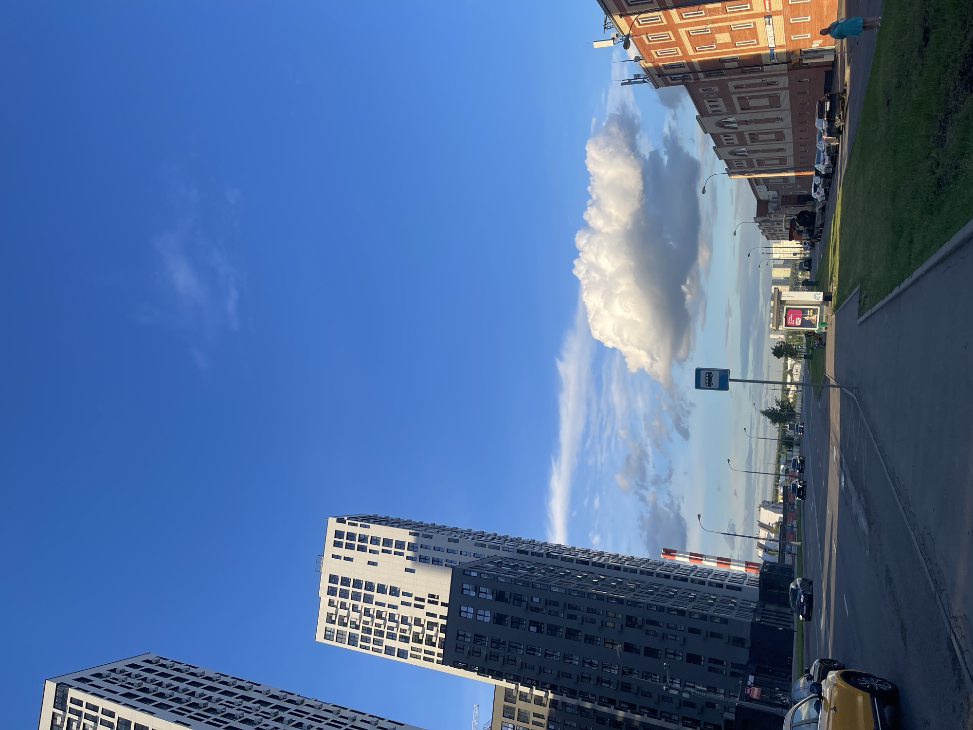 This media file captures an urban scene in Moscow, Russia, likely during the late afternoon or early evening, given the long shadows and the warm, golden light illuminating the buildings. The image is oriented vertically, with the sky dominating the upper portion. A large, fluffy white cloud is a prominent feature in the sky, contrasting with the deep blue.
The foreground features the sleek, modern facades of several high-rise apartment buildings. These buildings have a distinctive architectural style, characterized by repeating patterns of windows and balconies. One building is primarily white with a dark grey section, while another is entirely dark grey. In the lower right corner, parts of two cars are visible, one yellow and one dark-colored, parked on a street.
The middle ground shows a street lined with parked cars and streetlights. Further down the street, more buildings are visible, including a reddish-brown brick building with repeating window patterns, reminiscent of older architecture. A bus stop sign is clearly visible on a pole. The scene is one of everyday urban life, with no specific event depicted. The weather appears to be clear and pleasant, with the sun casting distinct shadows, indicating a sunny day with fair weather. The overall mood is calm and tranquil, highlighting the dynamic yet orderly nature of the modern cityscape. The tall buildings suggest a densely populated urban environment.