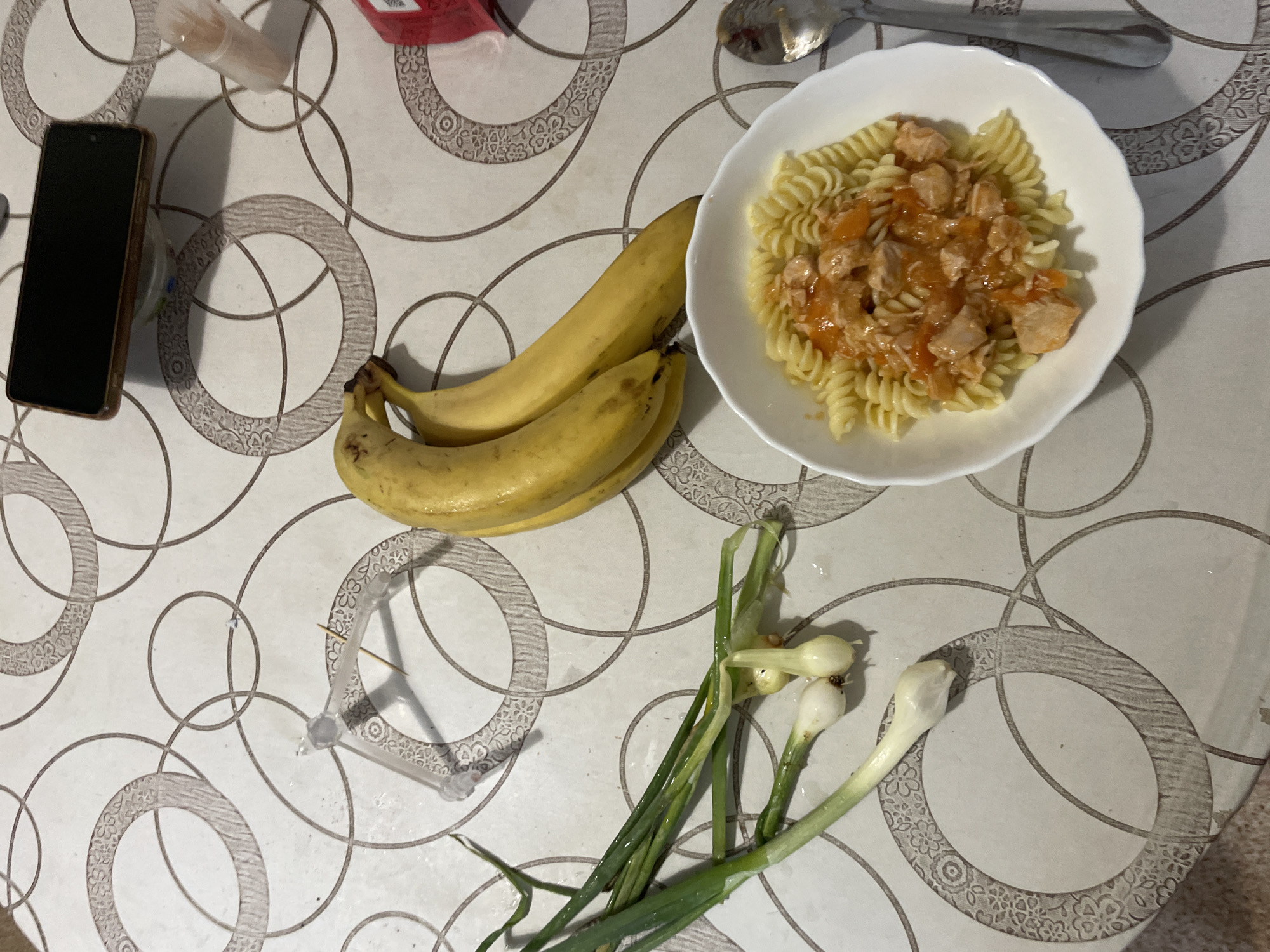 A top-down view shows a meal laid out on a patterned tablecloth. In the upper right quadrant, a white bowl filled with fusilli pasta and a topping of what appears to be chicken and tomato sauce is prominently displayed. To the left of the pasta, two ripe bananas are nestled together. Further to the left, a black smartphone lies face down. In the bottom right corner, a bunch of green onions, some with small bulbs, are arranged. A spoon rests to the right of the pasta bowl. The tablecloth features a repeating circular pattern with floral motifs within some of the circles. A small, translucent plastic object, possibly a toothpick holder with toothpicks, is visible near the top left of the image, partially casting a shadow. The lighting suggests it could be daytime, though there are no strong indicators of the time of day or weather. The setting appears to be a domestic environment, likely a dining table. There is no visible text in the image.