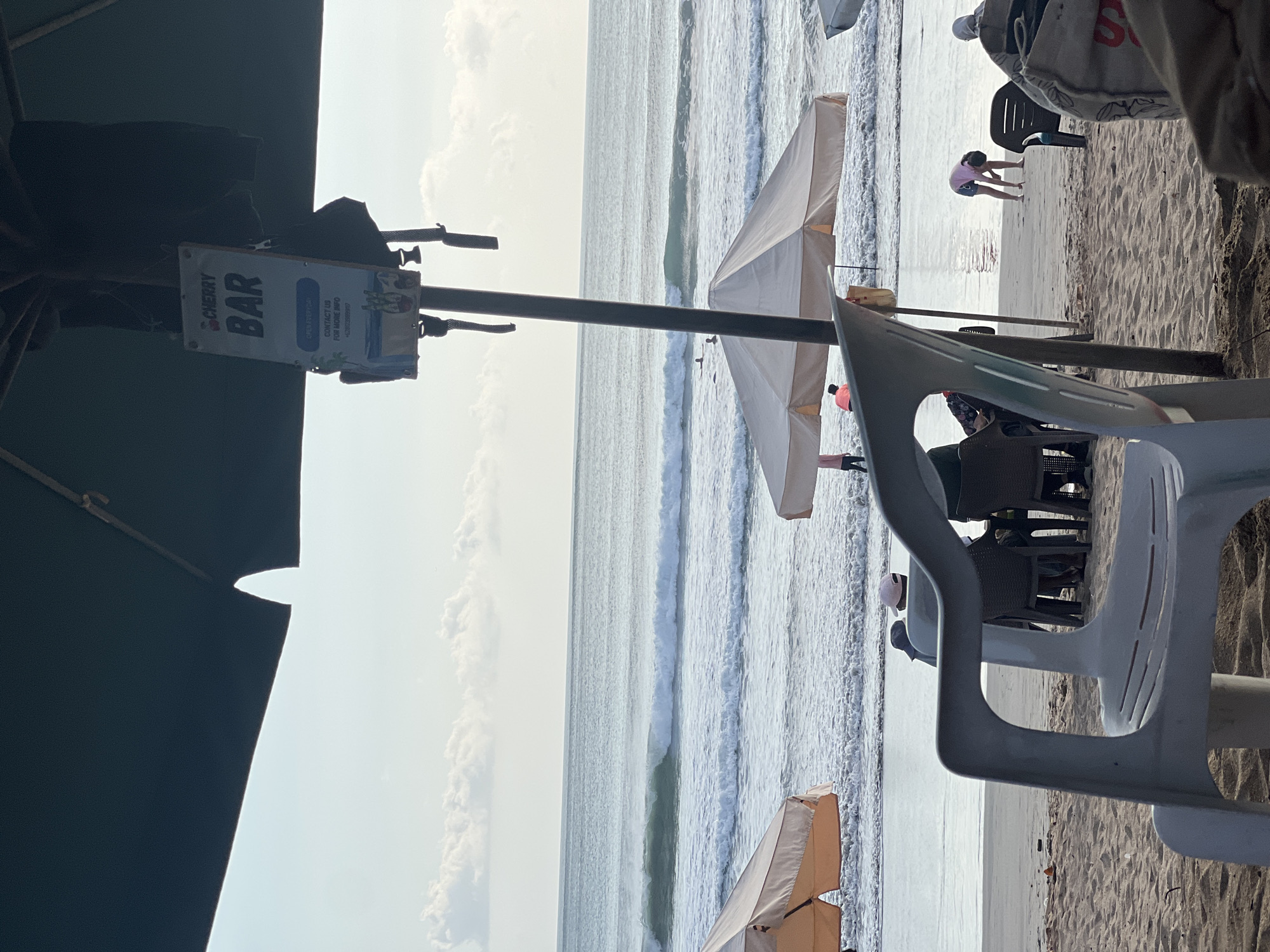This is a photograph taken from a beach in Kuta, Indonesia. In the foreground, a plastic chair is visible, and further back, several people are seated at tables under umbrellas. A young person in a pink shirt is bending over on the beach. The ocean is in the background with waves breaking and a few surfers in the water. A sign advertising "CHERRY BAR" hangs from an umbrella pole. The sky is light and cloudy.