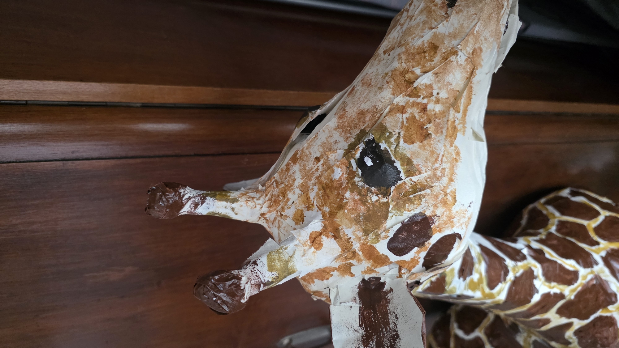 A close-up shows a handmade giraffe figure resting on or against a dark brown wooden surface, possibly a cabinet or table. The giraffe is constructed from paper or papier-mâché and painted with a brown and white pattern mimicking a giraffe's markings. The head and neck are prominent, with the details of the ears and muzzle being evident. The giraffe's body is partially visible in the background. The overall lighting is soft, creating shadows that enhance the textures of the giraffe figure and the wooden surface. The scene is sterile; no people, actions or interactions are occurring. The location is unknown.