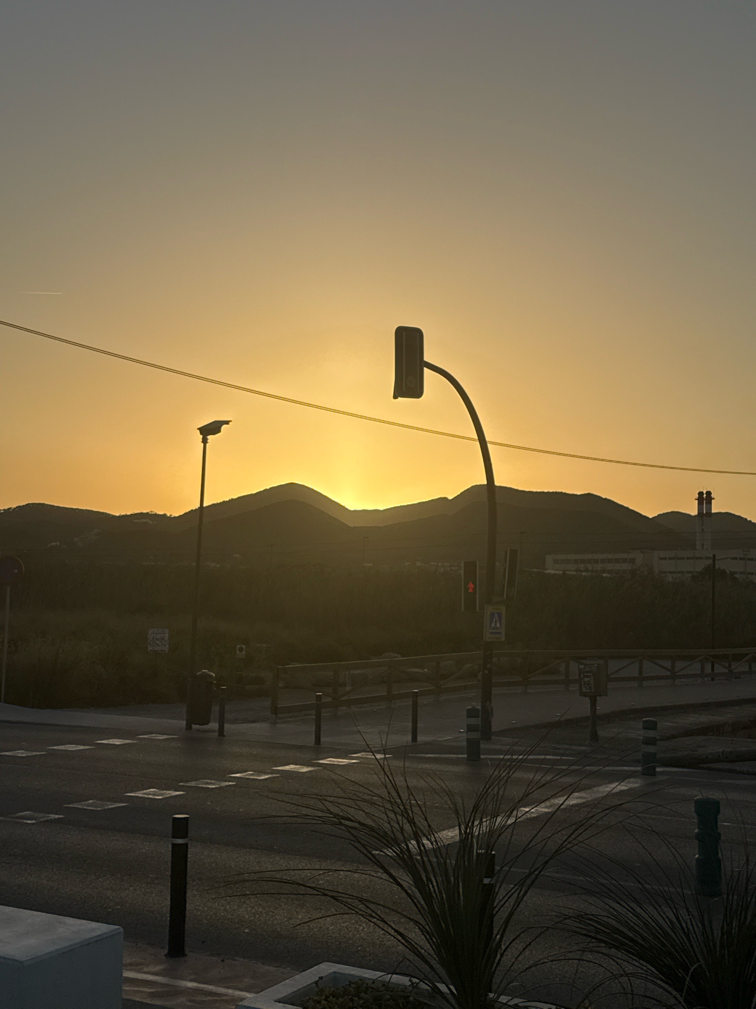 The image depicts a street scene at sunset in Ibiza, Spain. The sky is a warm, hazy yellow, with the sun low on the horizon, casting a soft glow over the landscape. In the foreground, a dark asphalt road features a pedestrian crossing marked by white squares. Several bollards, some striped, are strategically placed along the roadside.
A traffic light, with its signal facing the viewer, stands prominently on a curved pole. A pedestrian signal displaying a red stick figure is visible below the traffic light. Further back, a wooden fence delineates the edge of the road. Beyond the fence, silhouetted against the sunset, are rolling hills covered in dark vegetation, with industrial buildings and smokestacks visible in the distance. A street lamp, also silhouetted, stands to the left of the traffic light. The scene is devoid of any people or vehicles, creating a tranquil yet somewhat stark atmosphere typical of a developing area on the outskirts of a city. A power line stretches across the upper portion of the frame.