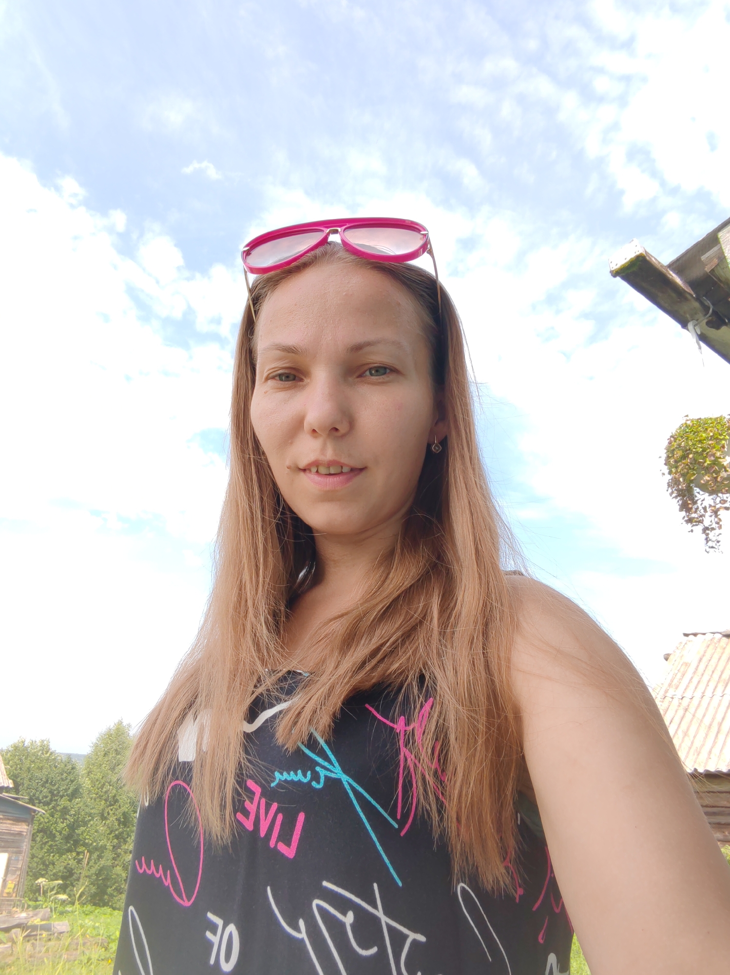The image is a selfie taken outdoors on a sunny day. A woman with long, light brown hair is centered in the frame, looking directly at the camera. She is wearing a black t-shirt with colorful graphic prints. Pink sunglasses are perched on top of her head. Her left ear has a small, dark earring. In the background, a bright blue sky with scattered white clouds is visible. To the right, a portion of a rustic wooden structure with a corrugated roof is present, and a plant hangs from it. To the left of the woman, lush green trees and a weathered wooden building are discernible. The overall setting suggests a rural or semi-rural environment, possibly in the city of Chukhlem, Russia, given the rustic architecture and natural landscape.