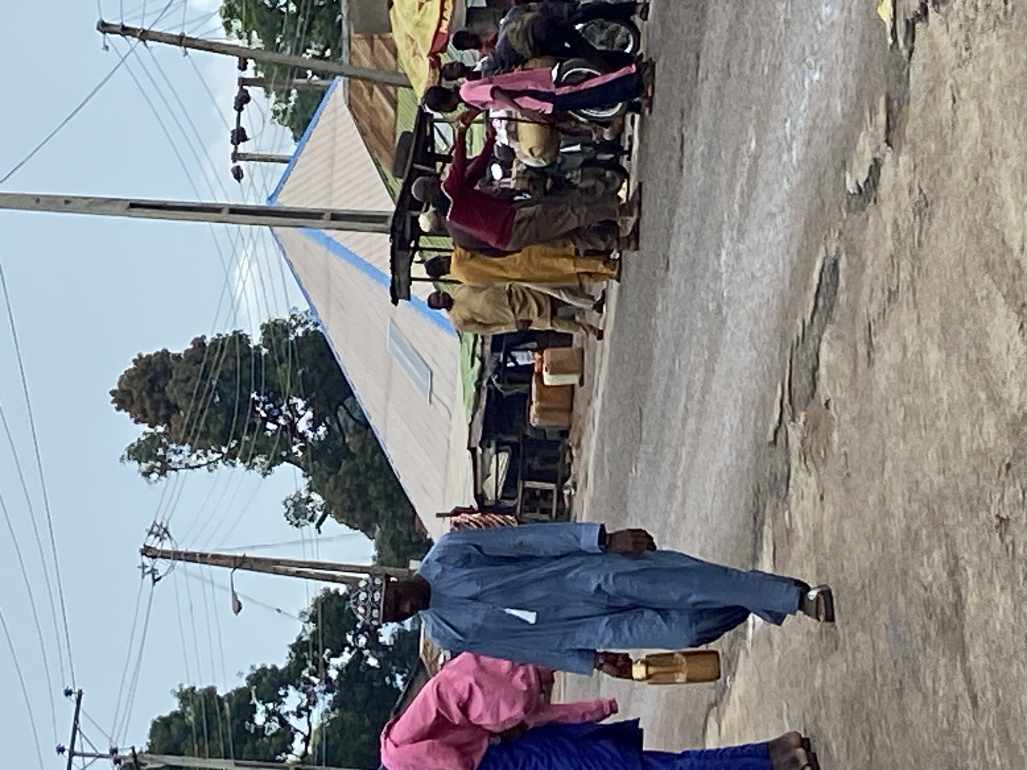 This image captures a street scene in Mararaba, Nigeria, under an overcast sky. The foreground features an uneven, worn asphalt road. On the left, a person in a blue lower garment and a pink top draped over their shoulder is partially visible, appearing barefoot. Adjacent to them, a man wearing a light blue traditional robe (agbada) and a white patterned skullcap walks towards the left, carrying a yellow jerrycan in his right hand. A white tassel hangs from his neck, and he wears sandals.
In the mid-ground, on the right side of the road, a group of approximately five to seven men is gathered around one or more motorcycles. One man, wearing a maroon long-sleeved top and brown trousers, extends his right arm towards another individual in a pink t-shirt and dark trousers, who stands near the motorcycle. Other men in tan or yellow traditional robes are also present within this group. One person in a dark garment is partially obscured, bending near the motorcycle. Several additional yellow jerrycans are visible on the ground behind this group, near makeshift stalls.
The background consists of various structures, including a prominent building with a light-colored corrugated metal pyramidal roof featuring a blue stripe and a rectangular solar panel. Further back, more rudimentary stalls or buildings with corrugated metal roofs are visible. Numerous wooden and concrete utility poles, some with attached streetlights, support a dense network of overhead power lines crisscrossing the sky. Trees with dark green foliage are scattered throughout the background. A yellow market umbrella is partially visible on the far right edge of the frame.